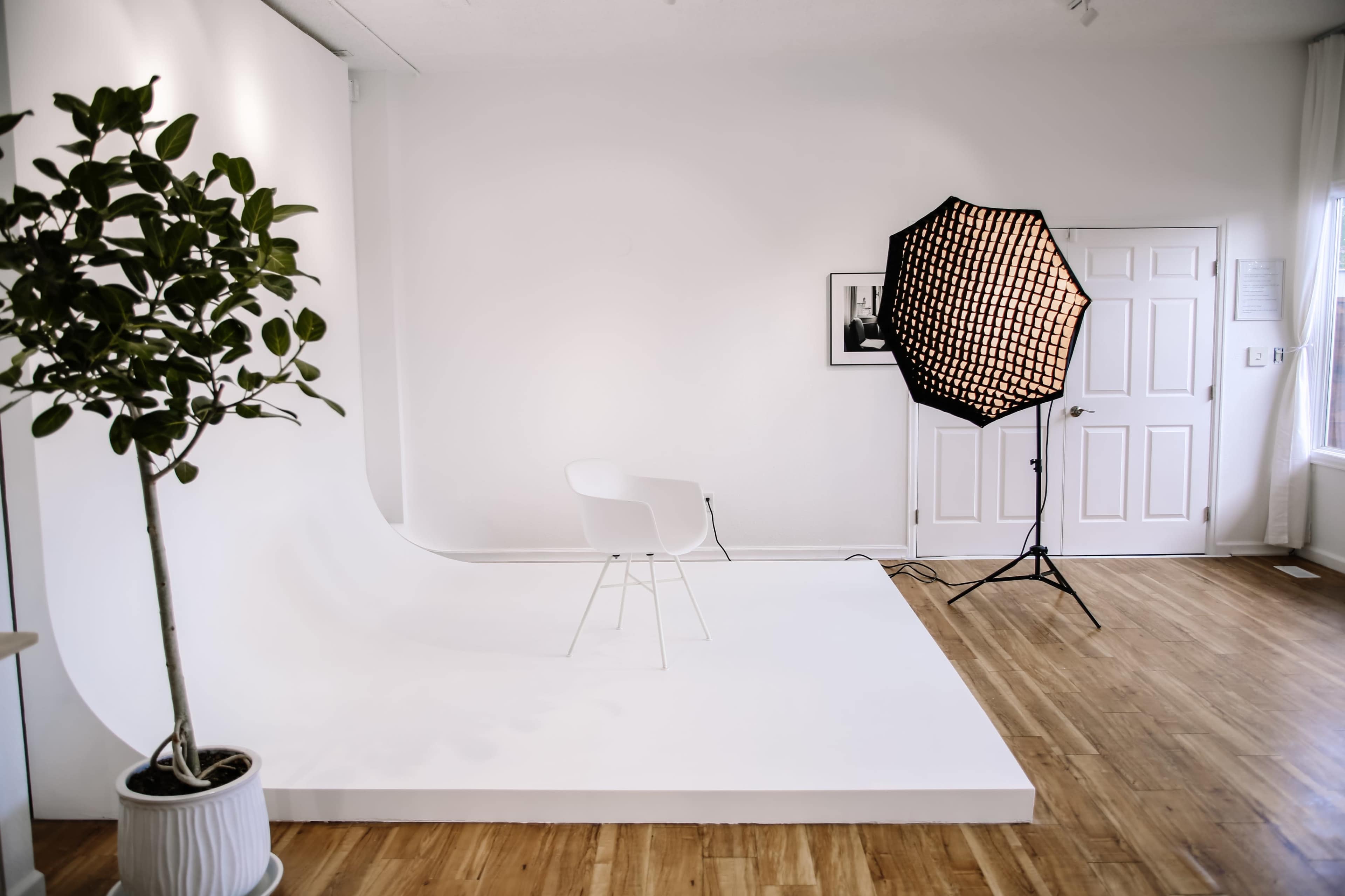 The image shows a minimalist photography studio with a white backdrop, a single white chair, and a large softbox light set up on the floor.