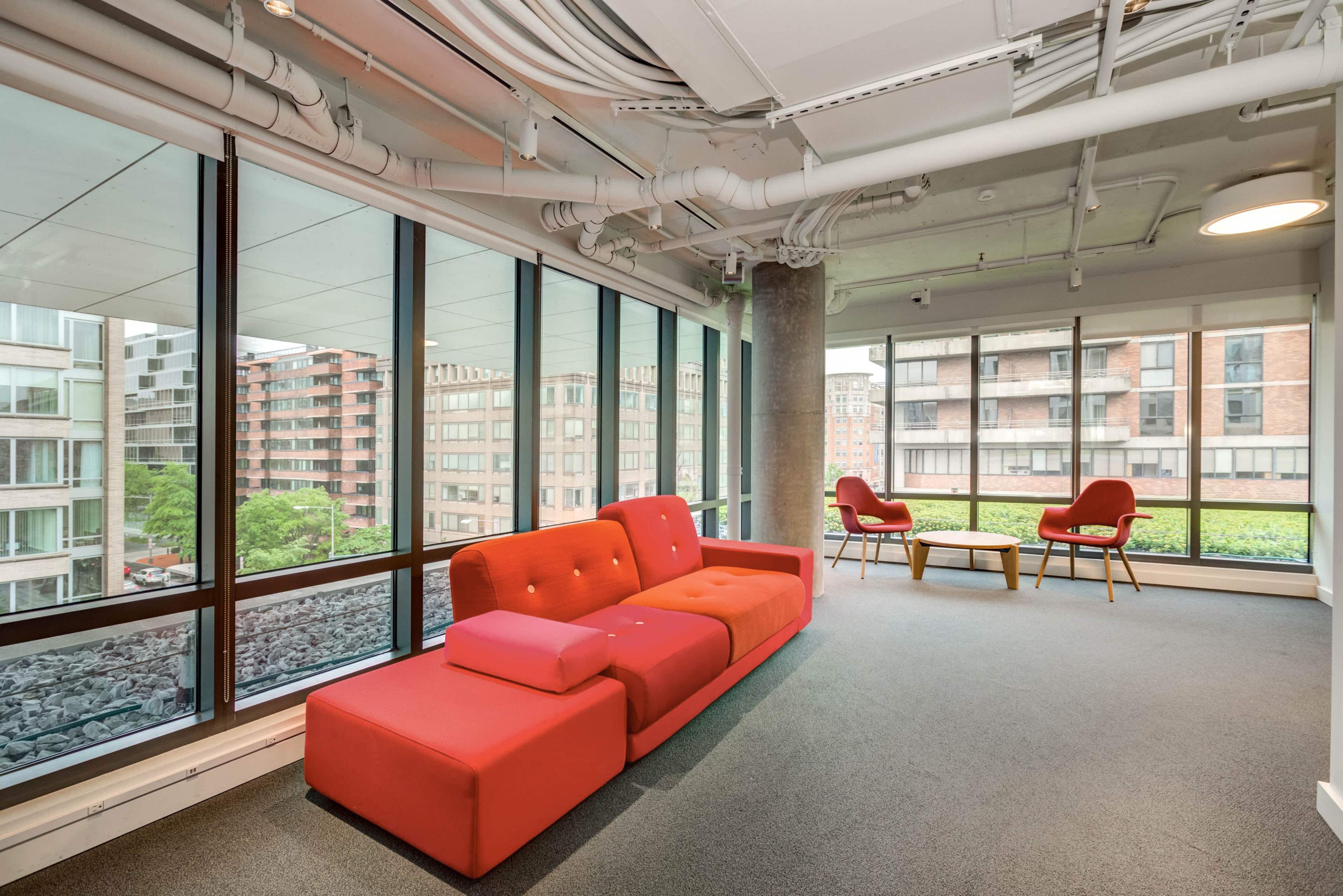The image shows a modern office space featuring a red sectional sofa, two red chairs, and large windows with a view of surrounding buildings.