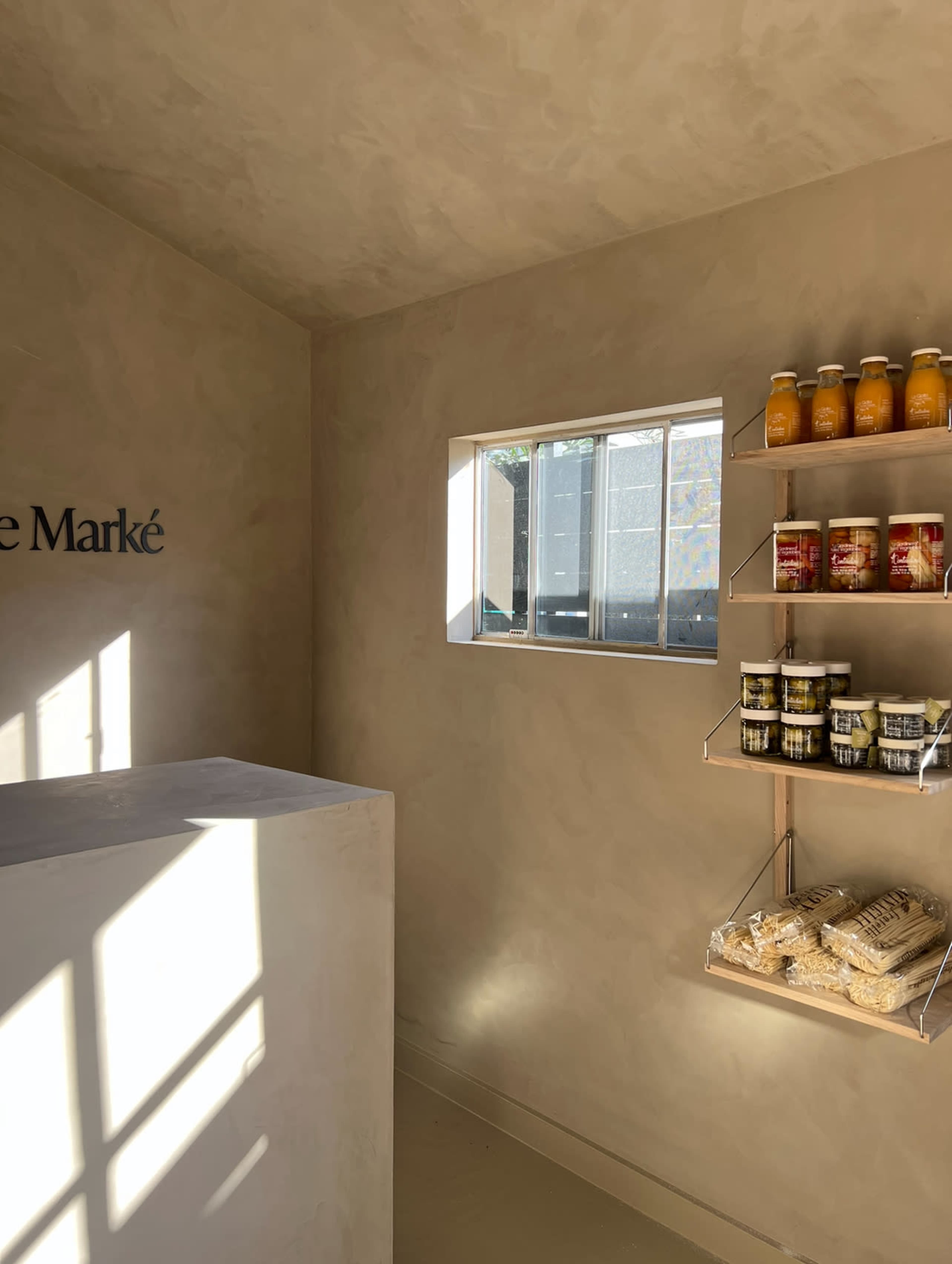 The image shows a minimalistic interior space with a counter on the left and shelves displaying jars and pasta on the right, illuminated by natural light from a window.
