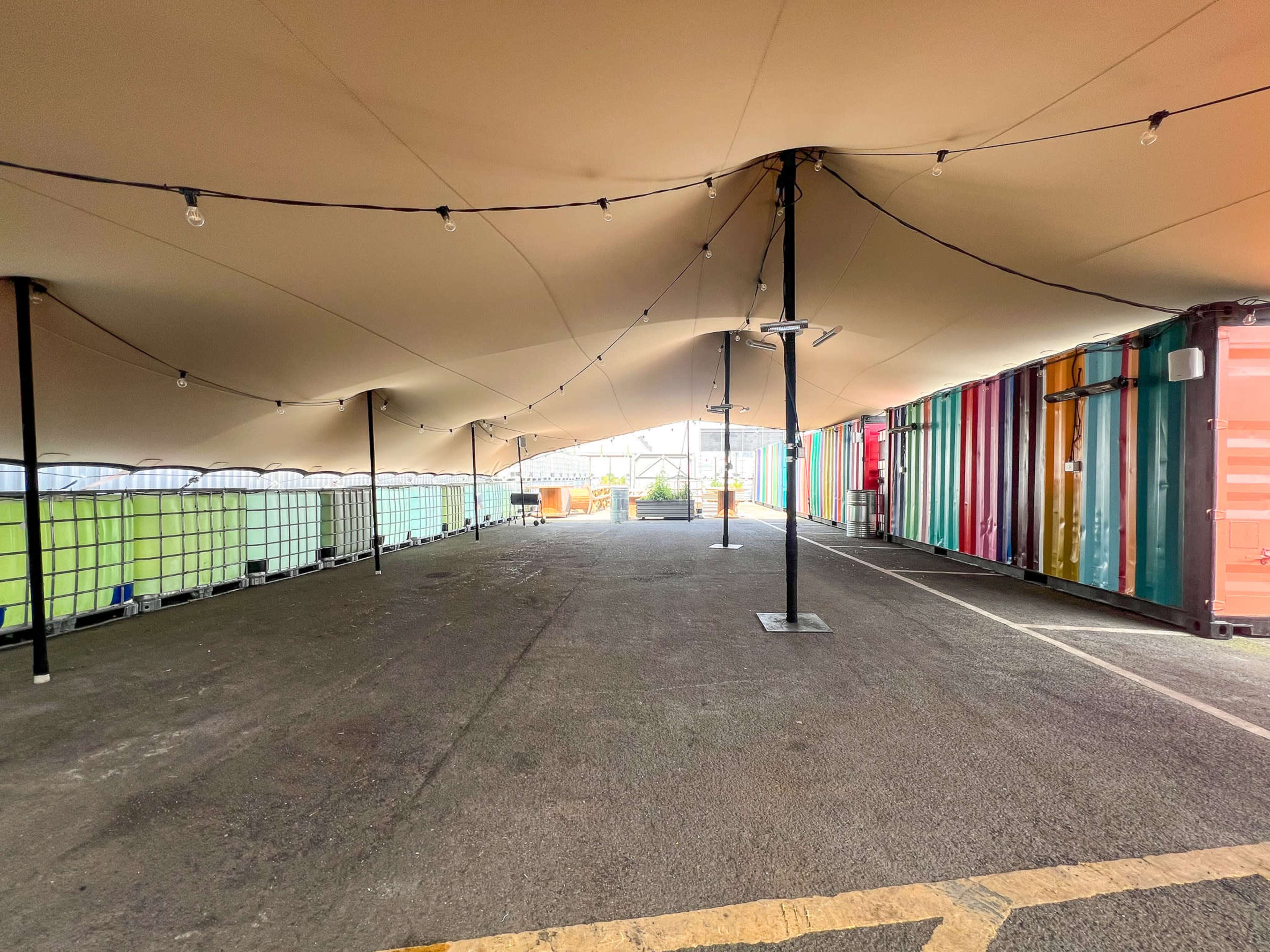 An outdoor area covered by a large tent, lined with shipping containers on one side and illuminated by string lights.