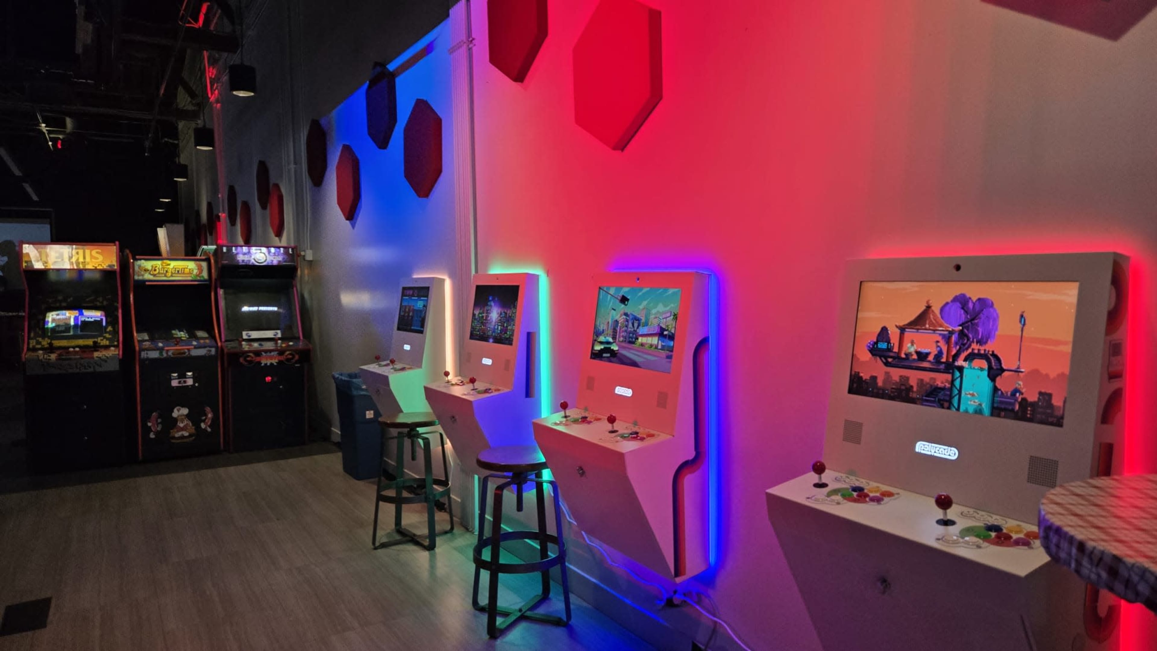 The image shows a row of colorful arcade game machines against a wall, with stools in front and traditional arcade cabinets visible in the background.