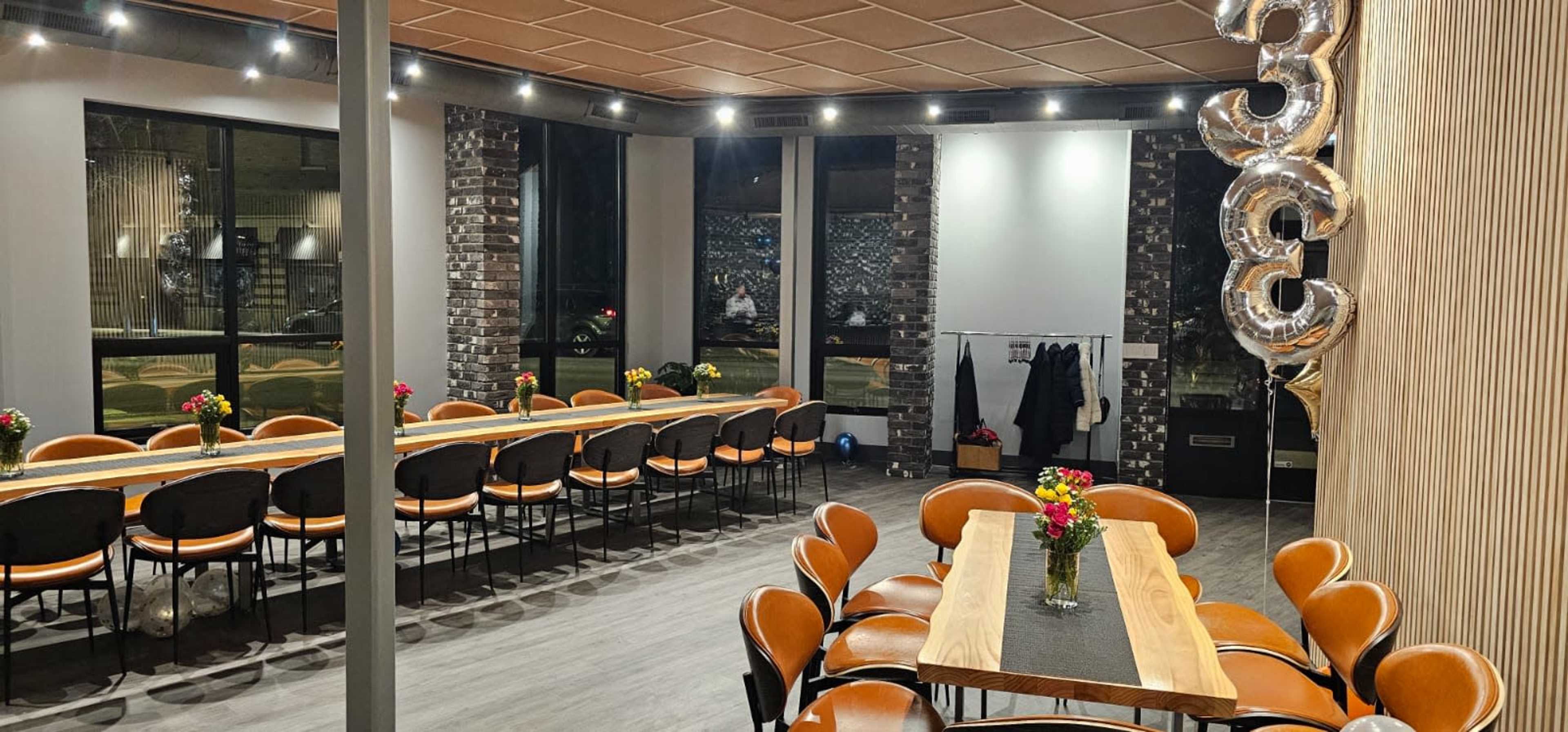 A spacious dining area features long wooden tables adorned with flower vases, surrounded by black chairs under warm lighting.