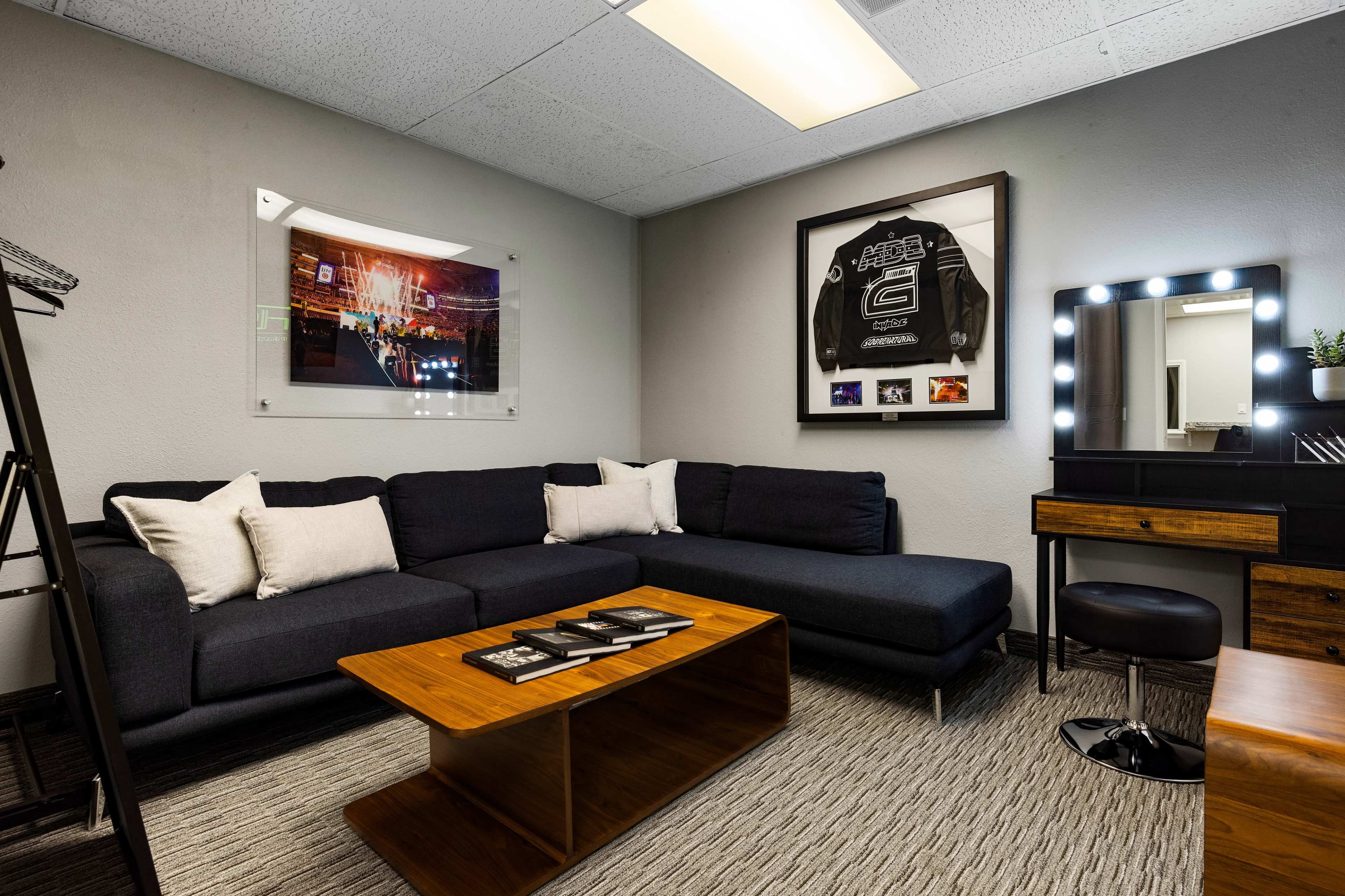 A modern lounge area with a dark sofa, a wooden coffee table, and a vanity mirror illuminated by lights, featuring framed artwork and a racing jacket on the wall.