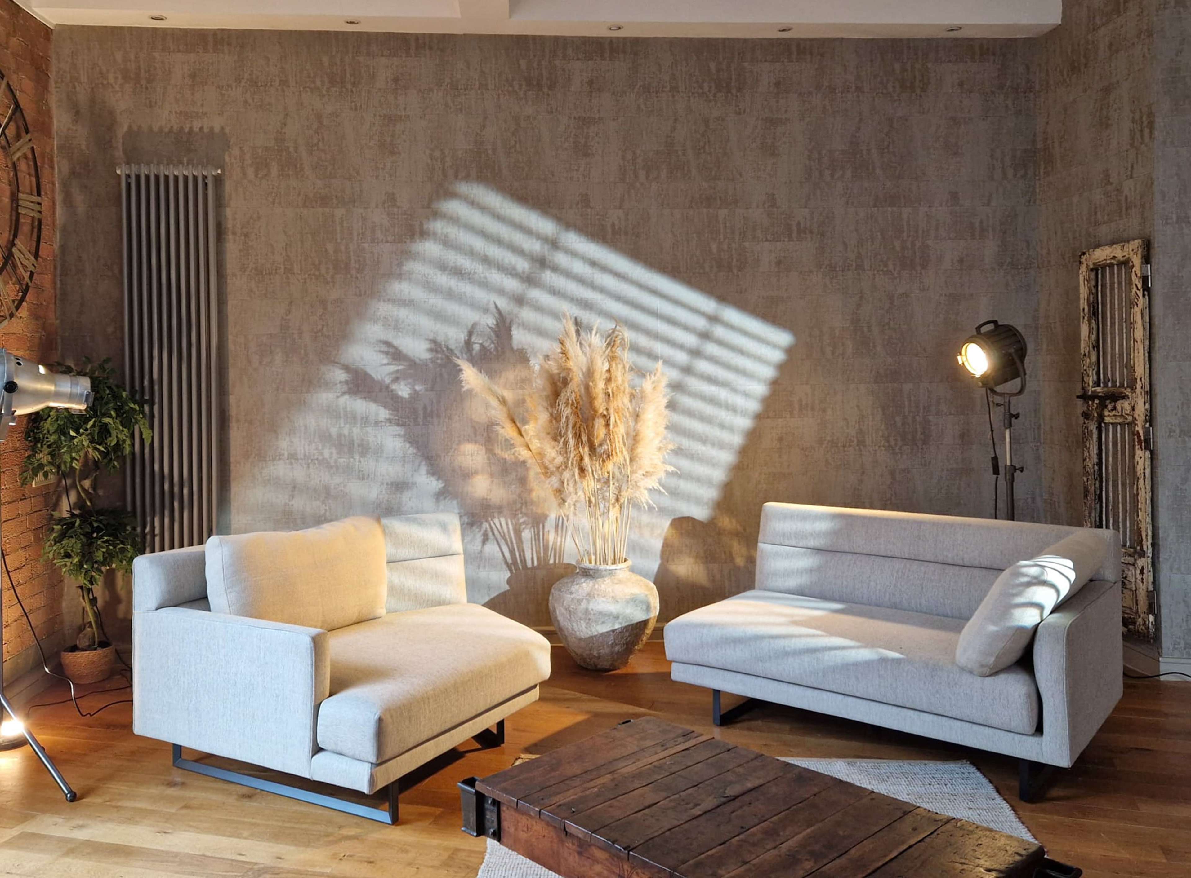 A well-lit living room features two light gray sofas positioned around a wooden coffee table, with a decorative plant arrangement in a large vase and a spotlight illuminating the scene.