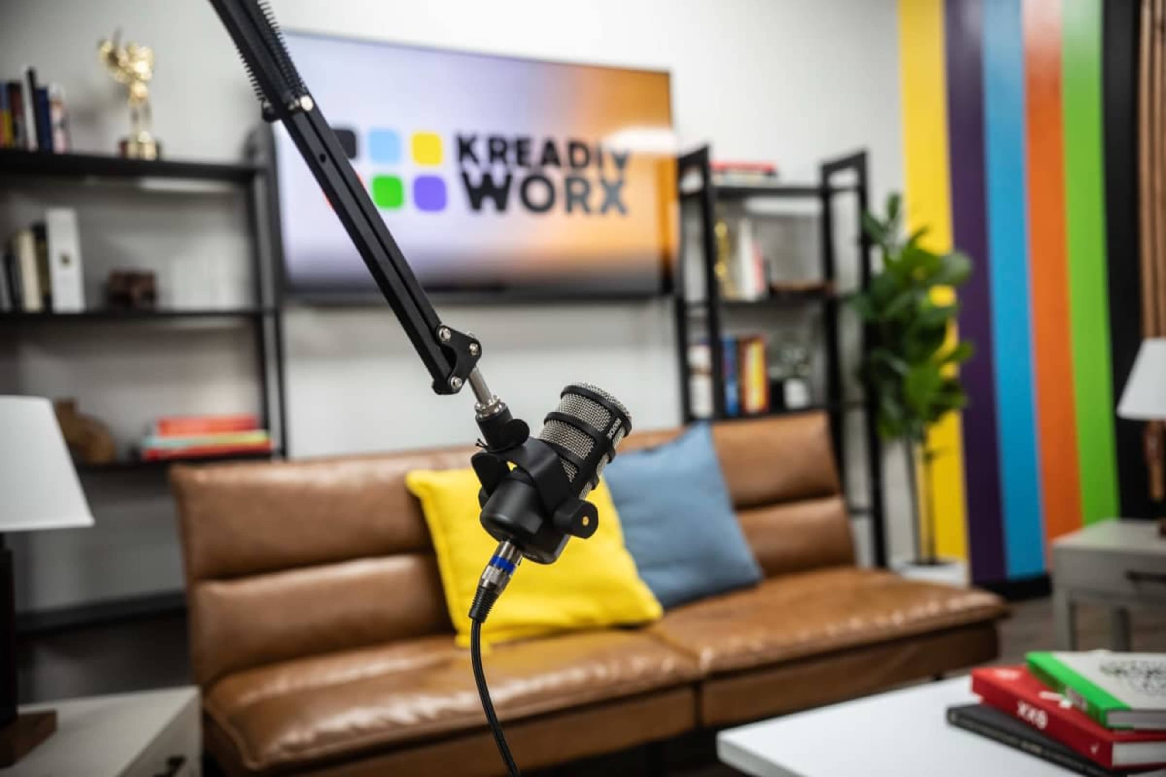 The image shows a microphone on a boom arm in a modern studio setting with a colorful striped wall and a logo for "Kready Worx" displayed on a screen in the background.