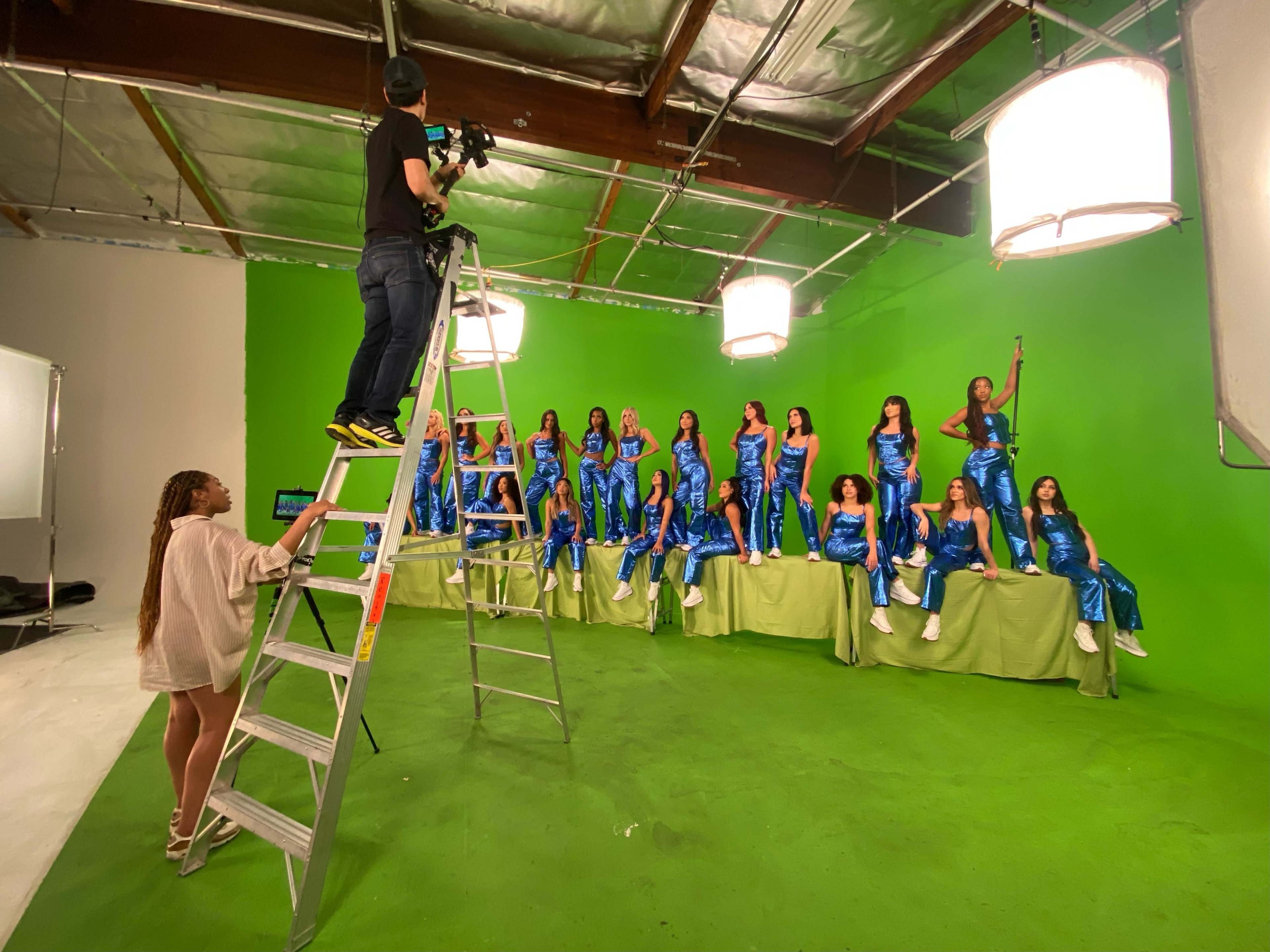 A film crew captures a group of models dressed in blue outfits while posing on a green backdrop.