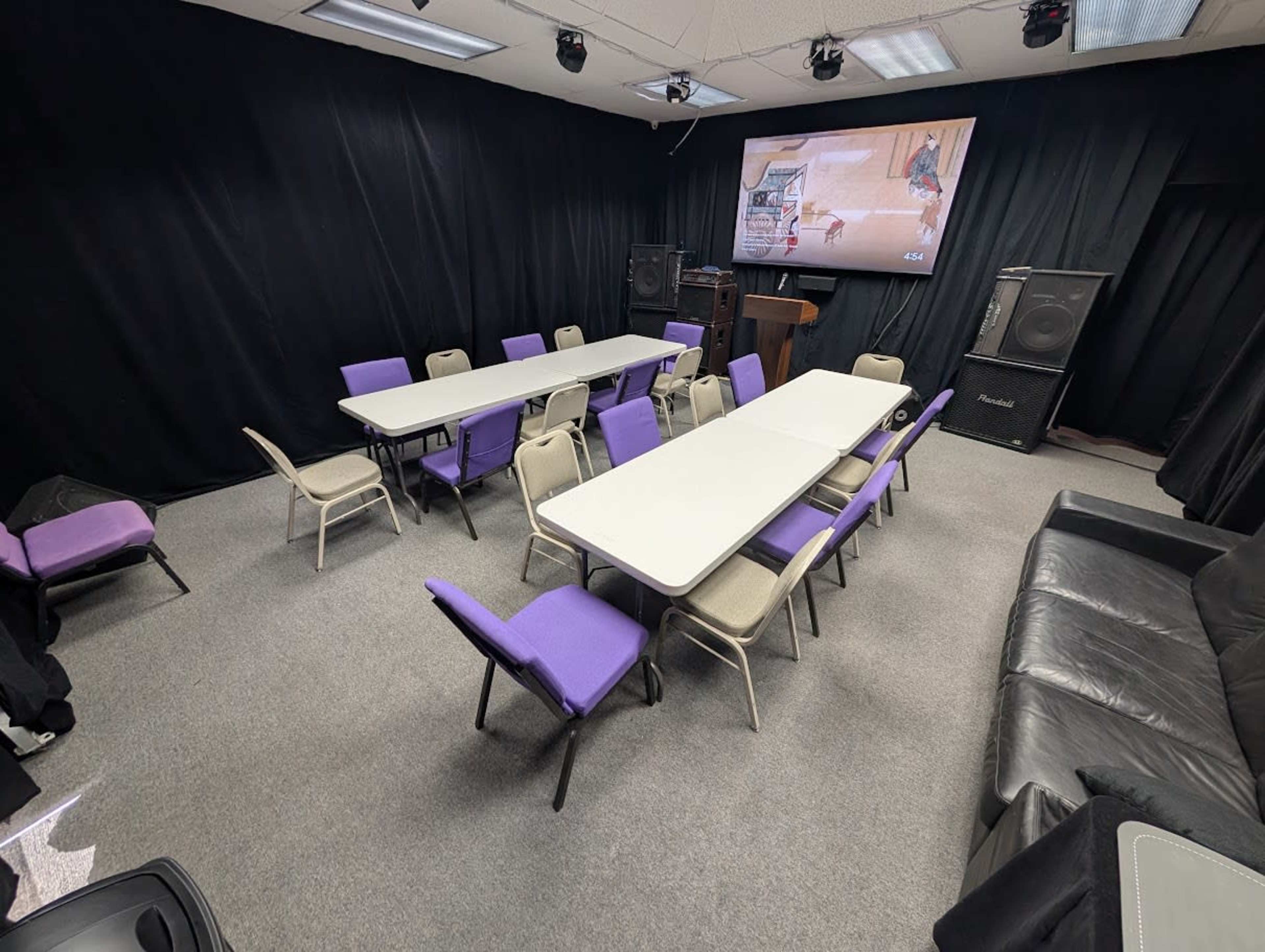 The image shows a meeting room with multiple tables arranged in a U-shape, surrounded by chairs, and features a large screen at the front and audio equipment on the sides.