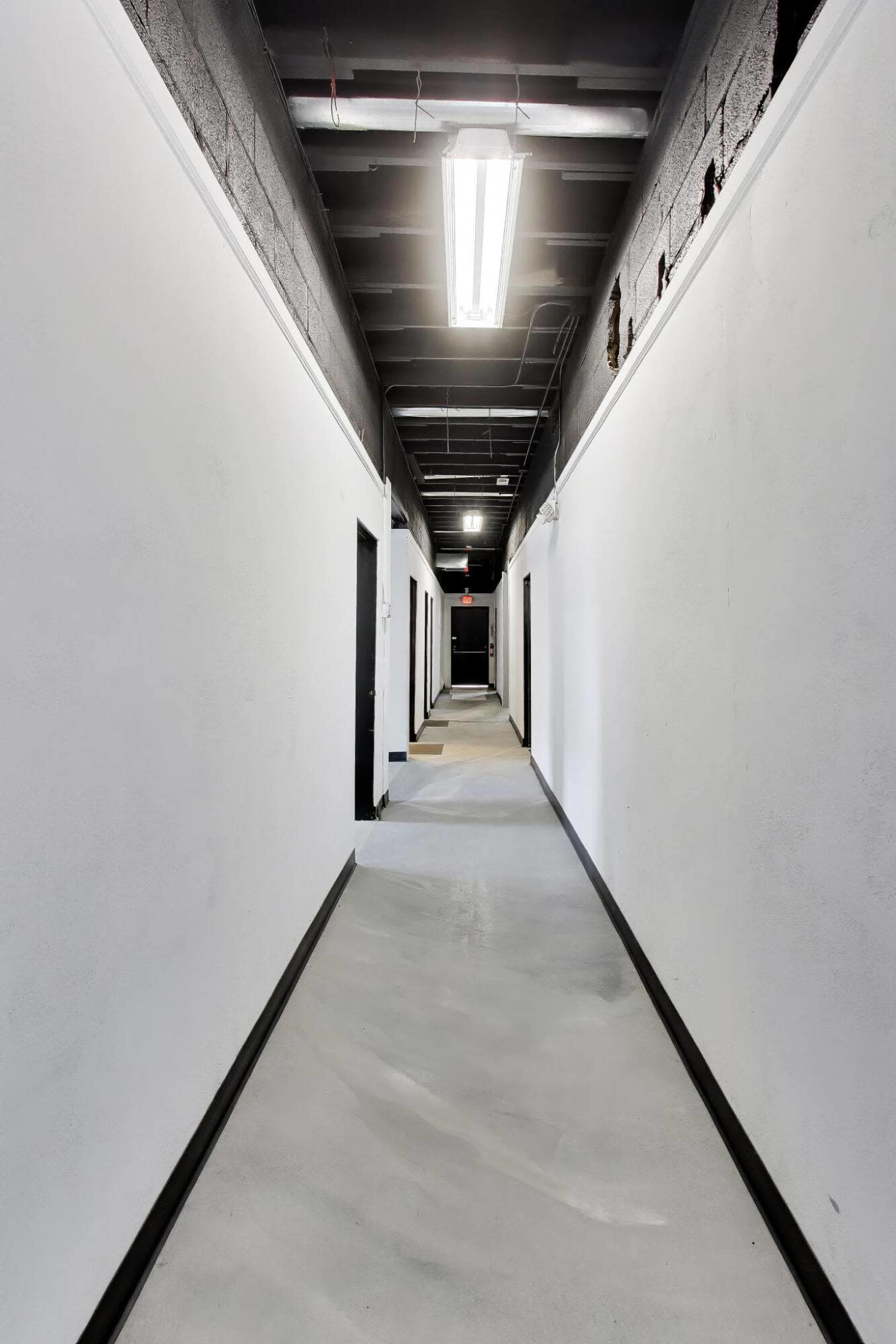 The image shows a long, empty hallway with white walls and several closed doors on either side.