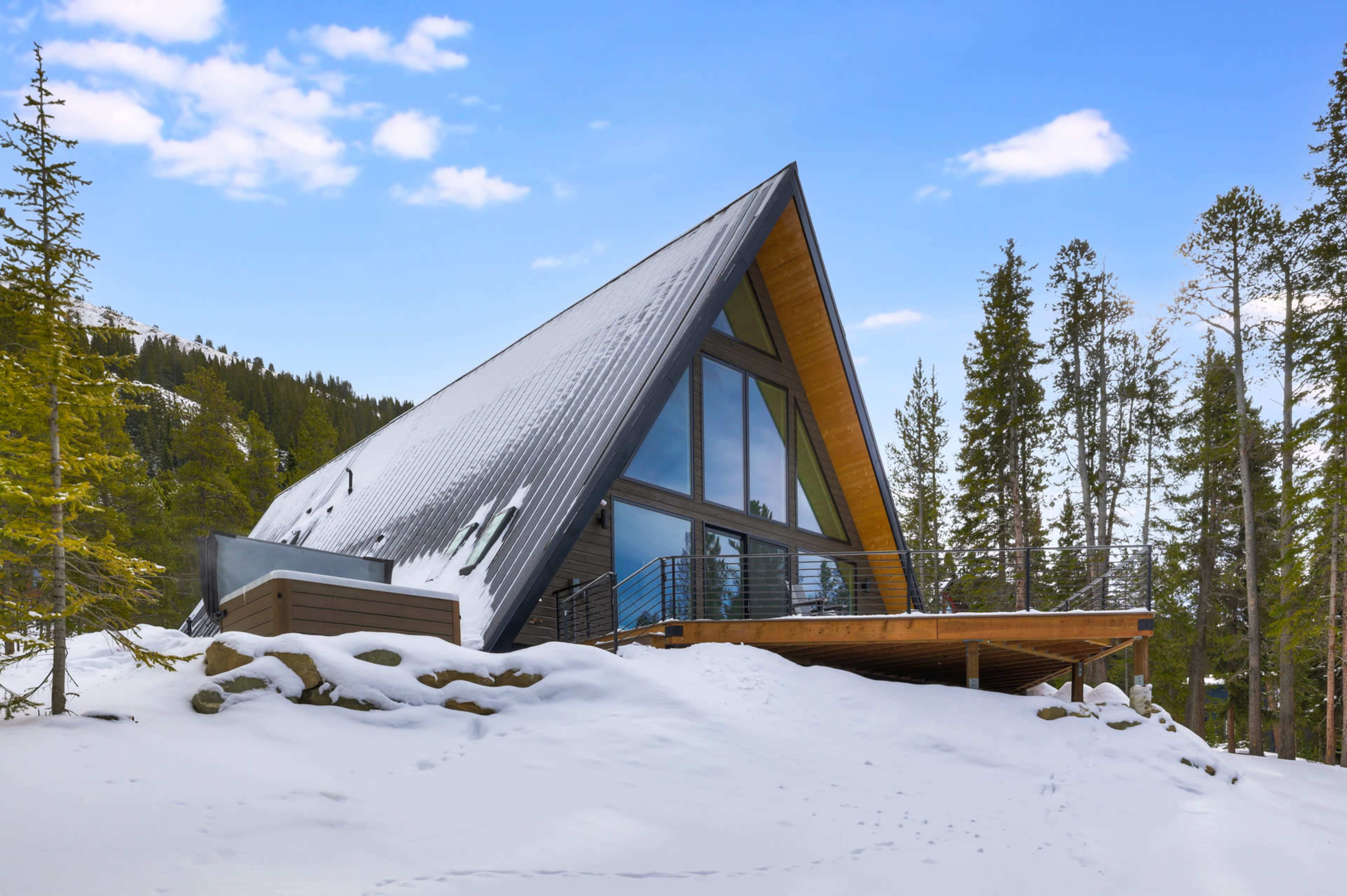 A modern A-frame cabin with large windows sits on a snowy landscape surrounded by trees.