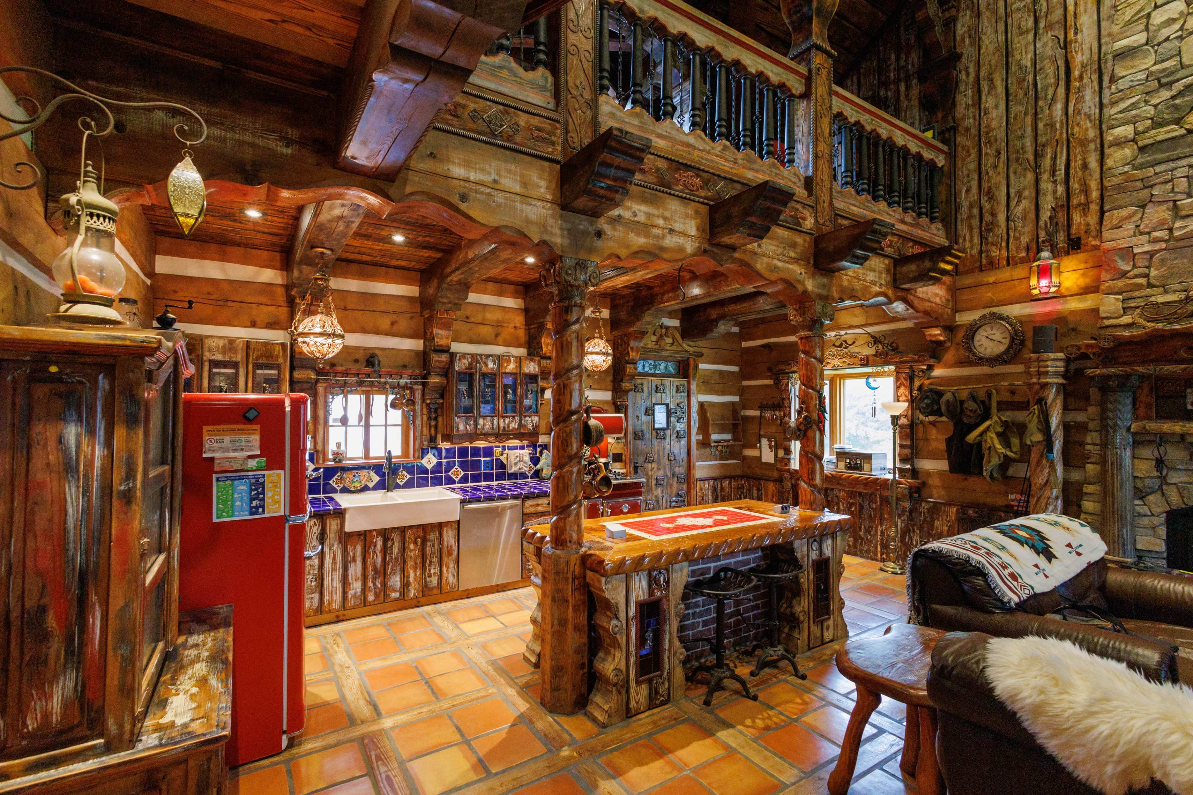 The image shows a rustic wooden kitchen with a red refrigerator, stone accents, and ornate wooden beams in a cozy cabin setting.