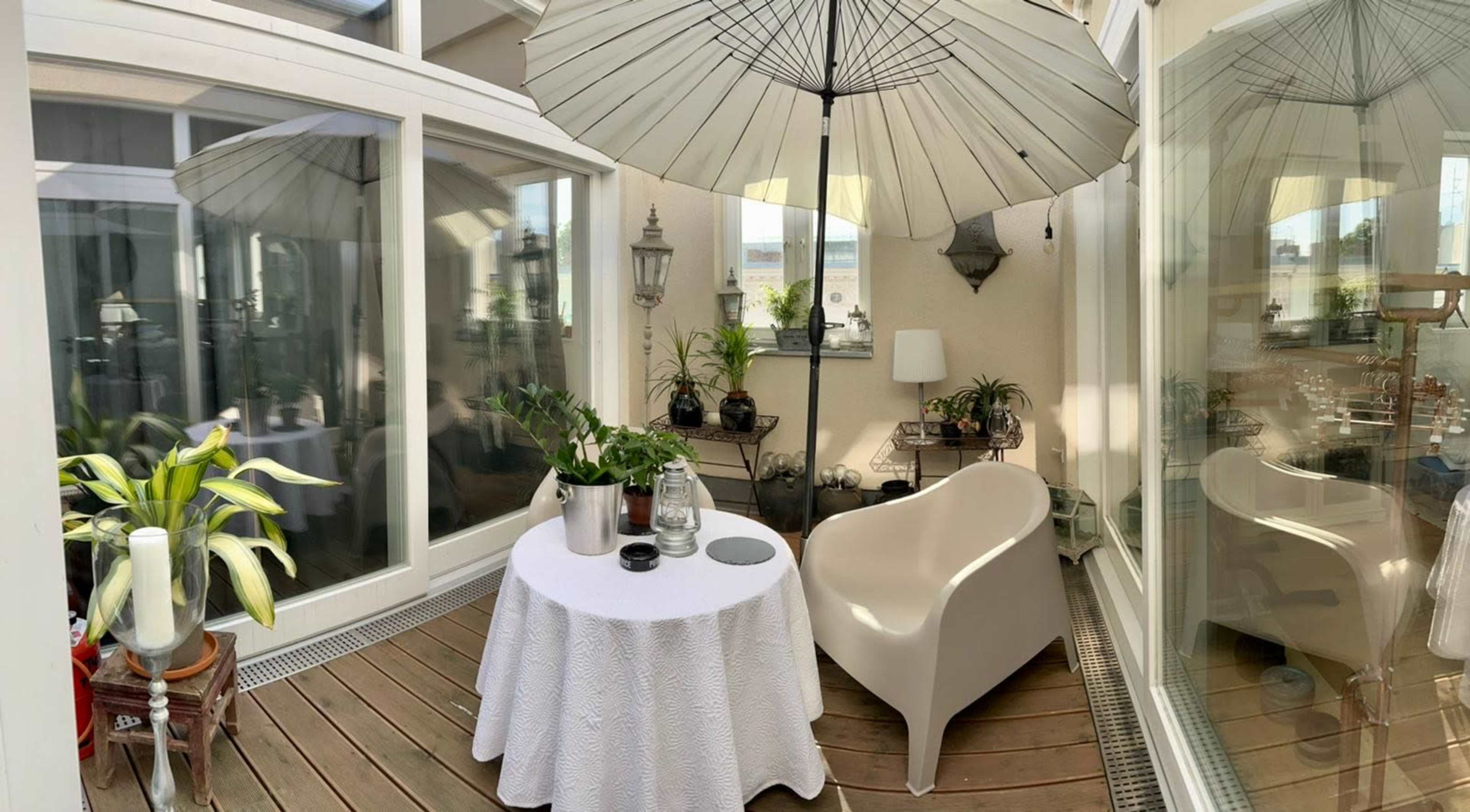 A small, enclosed balcony features a white table with a tablecloth, a chair, an umbrella, potted plants, and large windows.