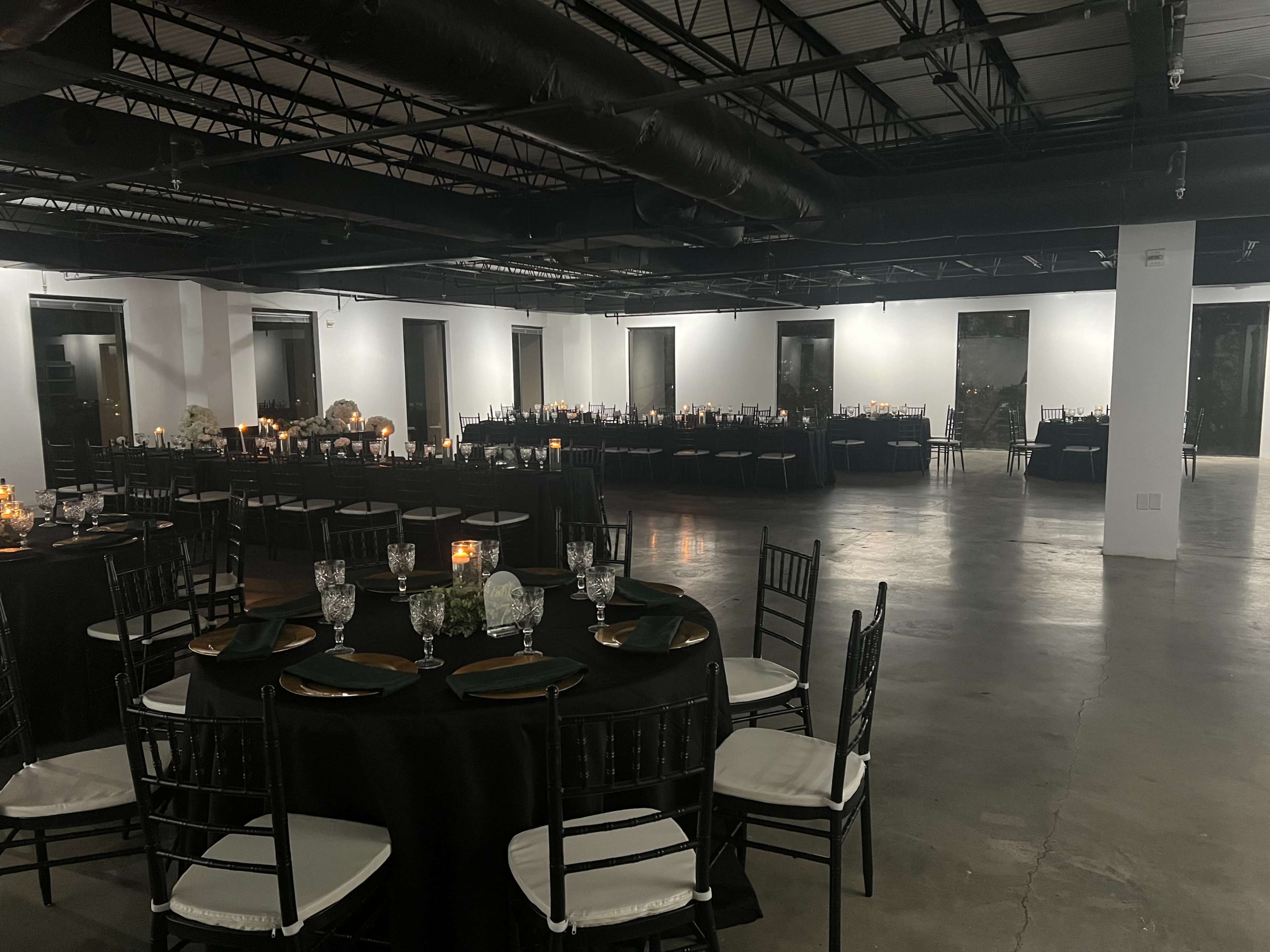 A spacious event venue set up for a formal gathering, featuring multiple round tables with black tablecloths and chairs arranged around them, illuminated by candlelight.