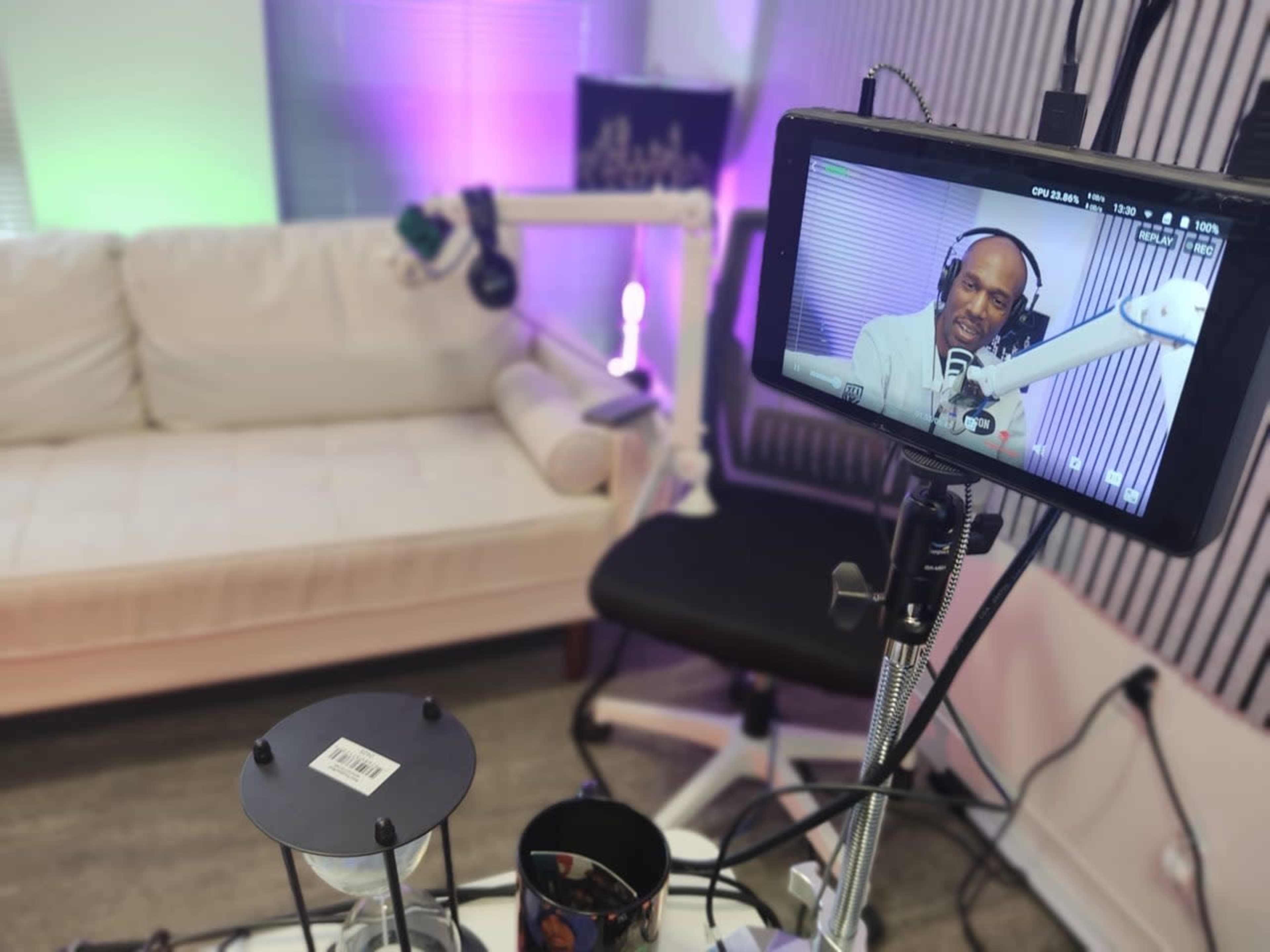 The image shows a recording setup with a tablet displaying a person speaking, a microphone on a stand, a cozy white couch in the background, and a black chair nearby.