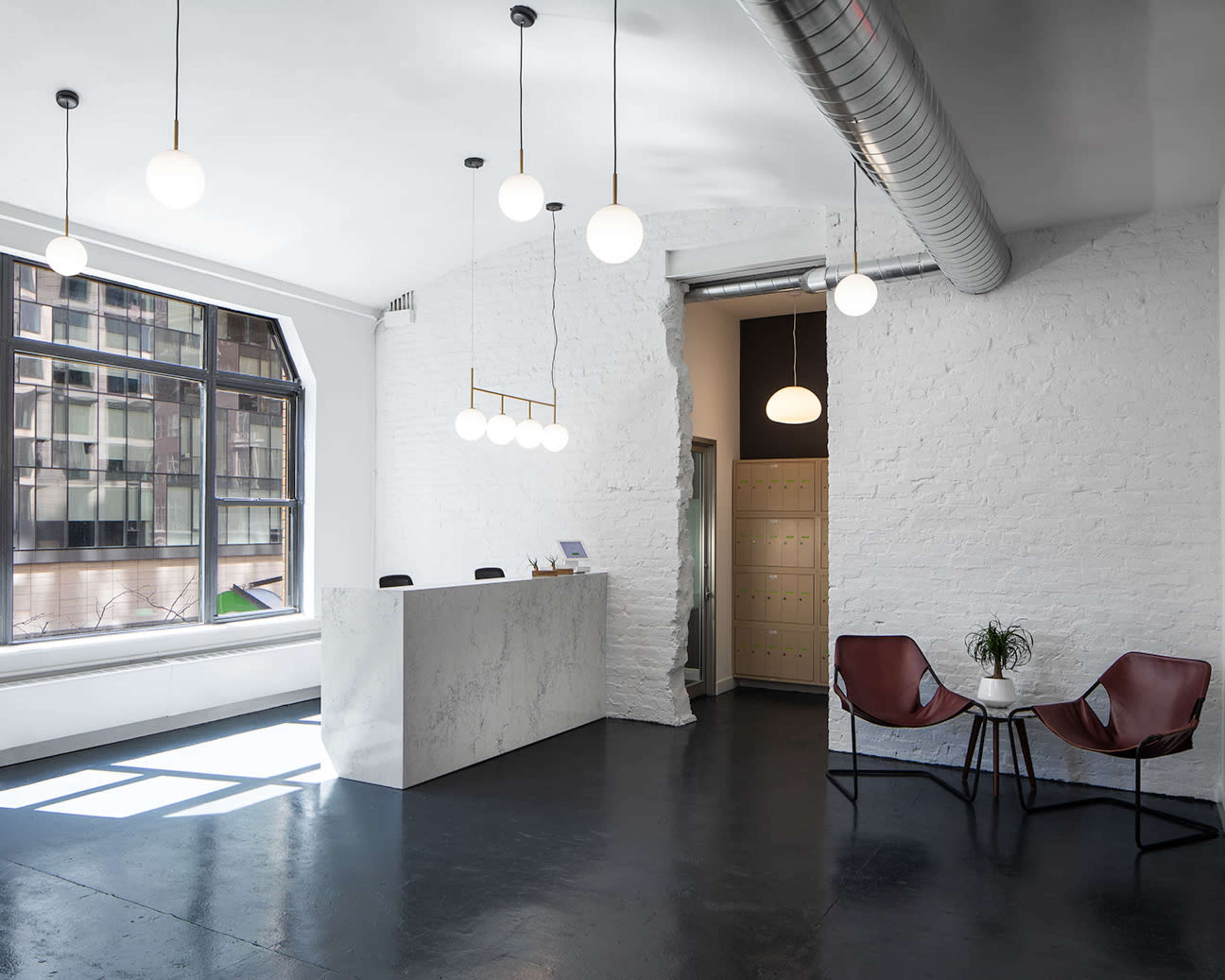 A modern reception area with a white marble desk, hanging globe lights, and a seating space featuring two brown chairs and a small plant.