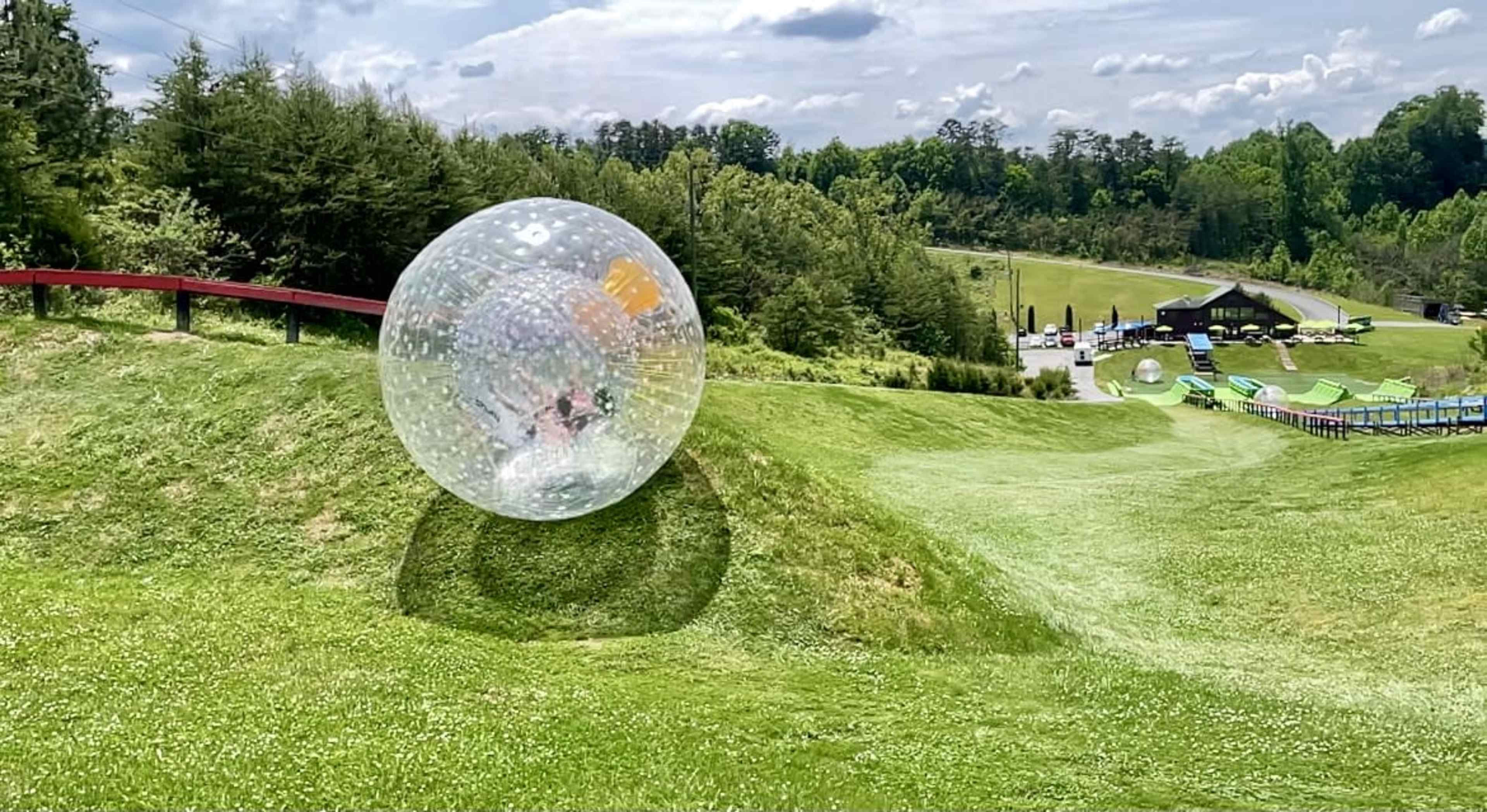 A large transparent inflatable sphere rolls down a grassy hill at a recreational park with trees and a building in the background.