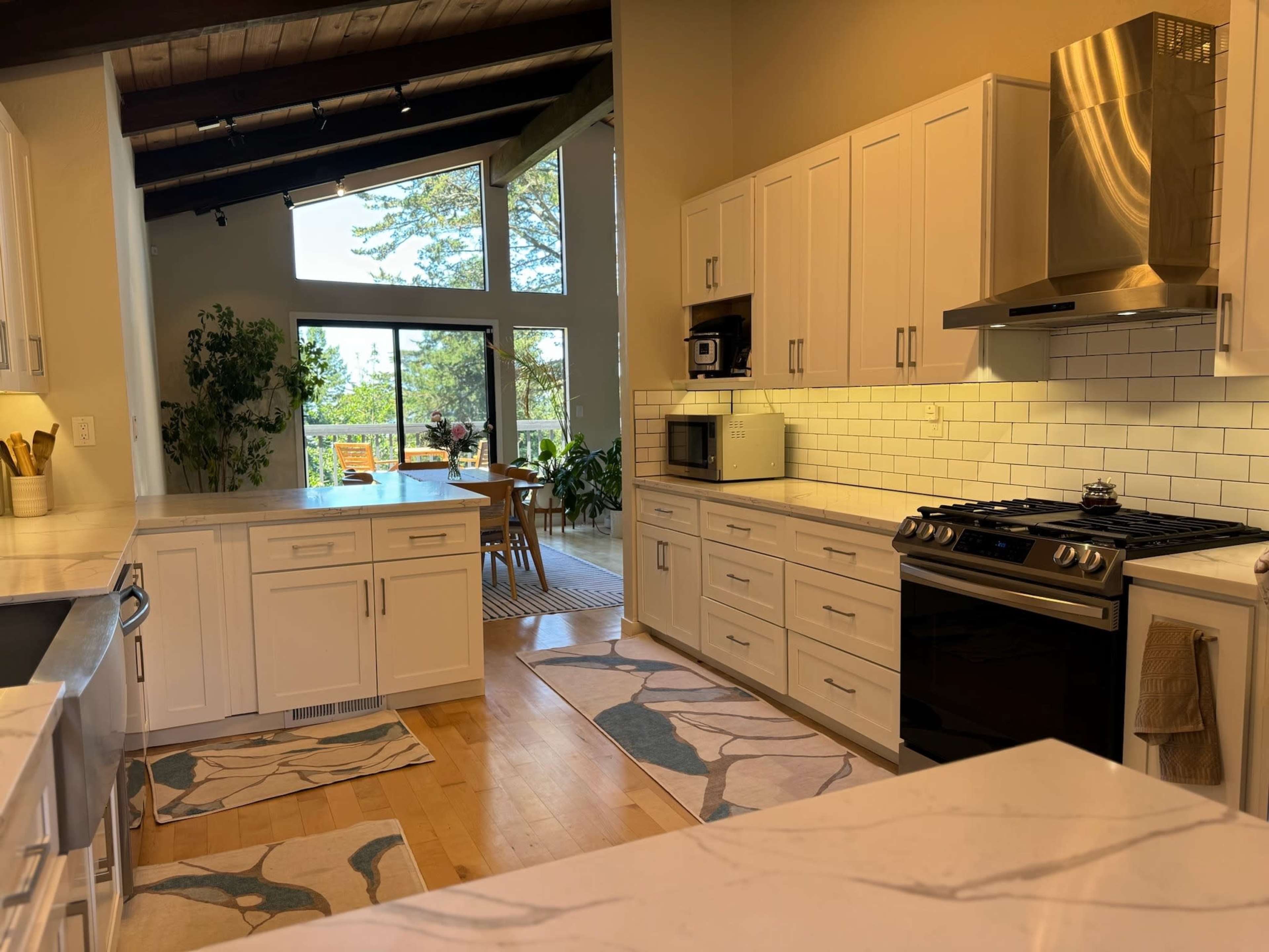 A modern kitchen features white cabinets, a gas stove, and large windows overlooking a dining area filled with plants.