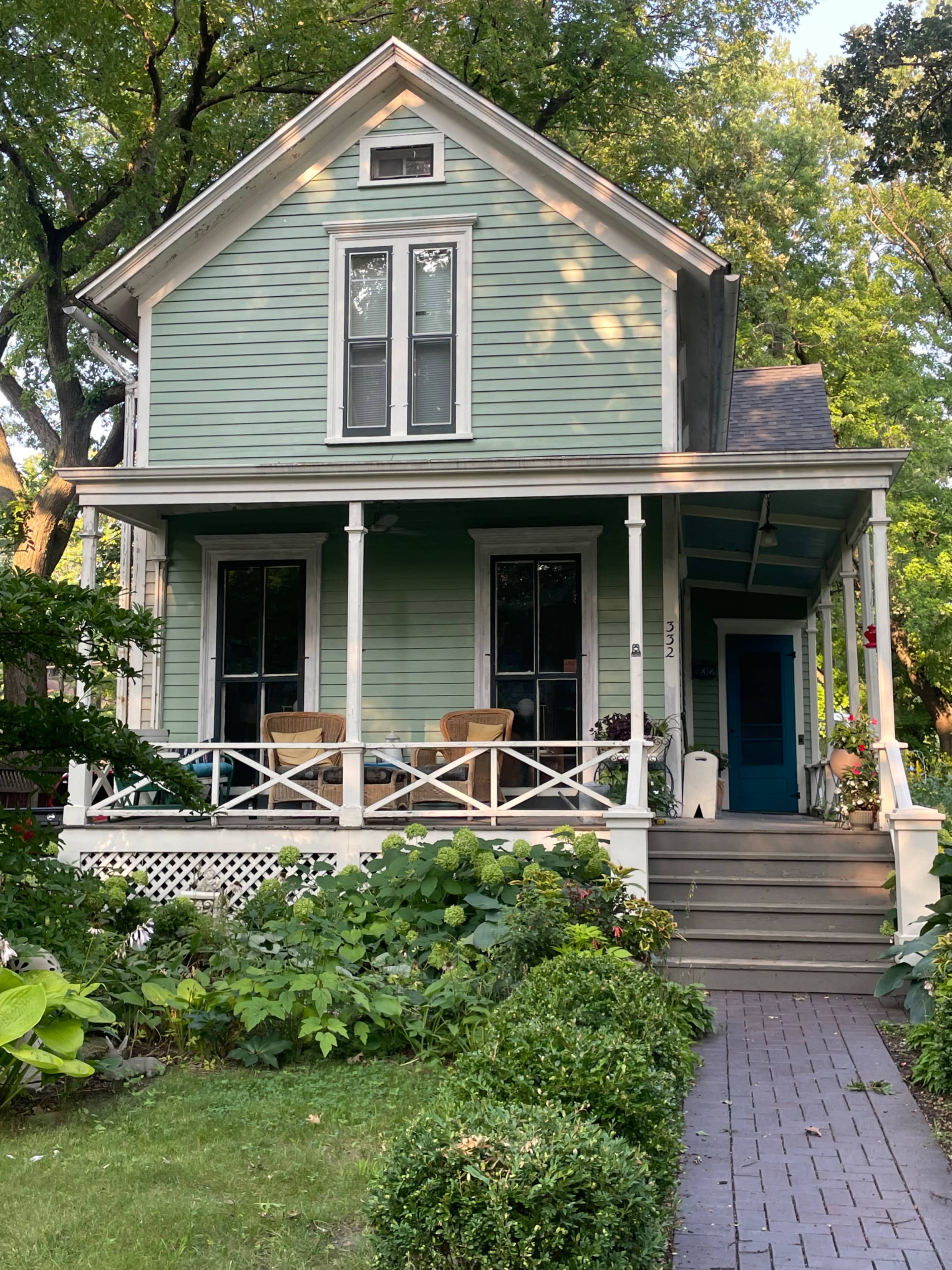 A green, two-story house with a front porch and a neatly landscaped yard features a brick walkway leading to the entrance.