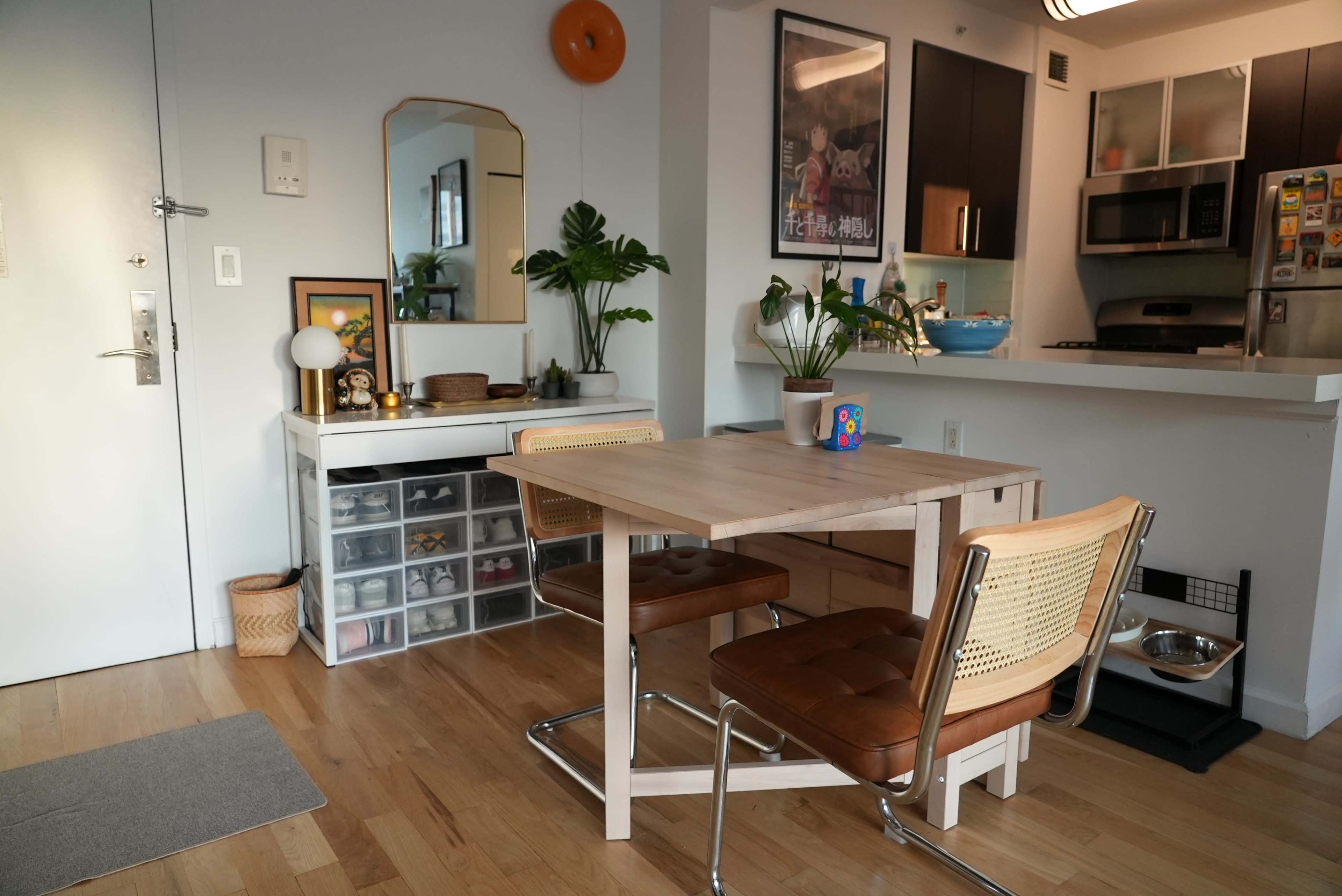 A modern dining area features a wooden table with two chairs, a nearby mirror, and a kitchen partially visible in the background.