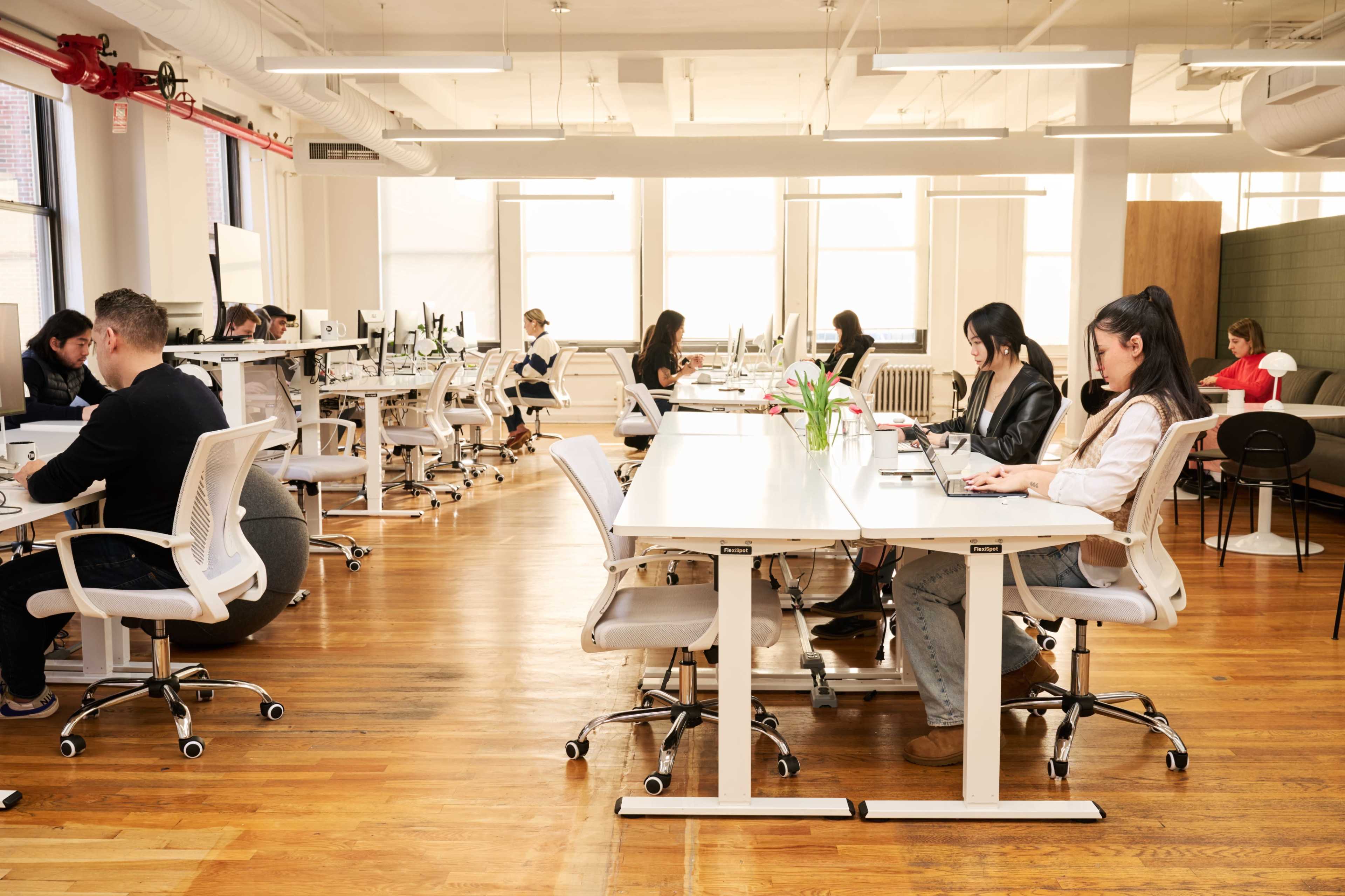 The image shows a modern office space with several people working at desks, surrounded by large windows and hardwood flooring.