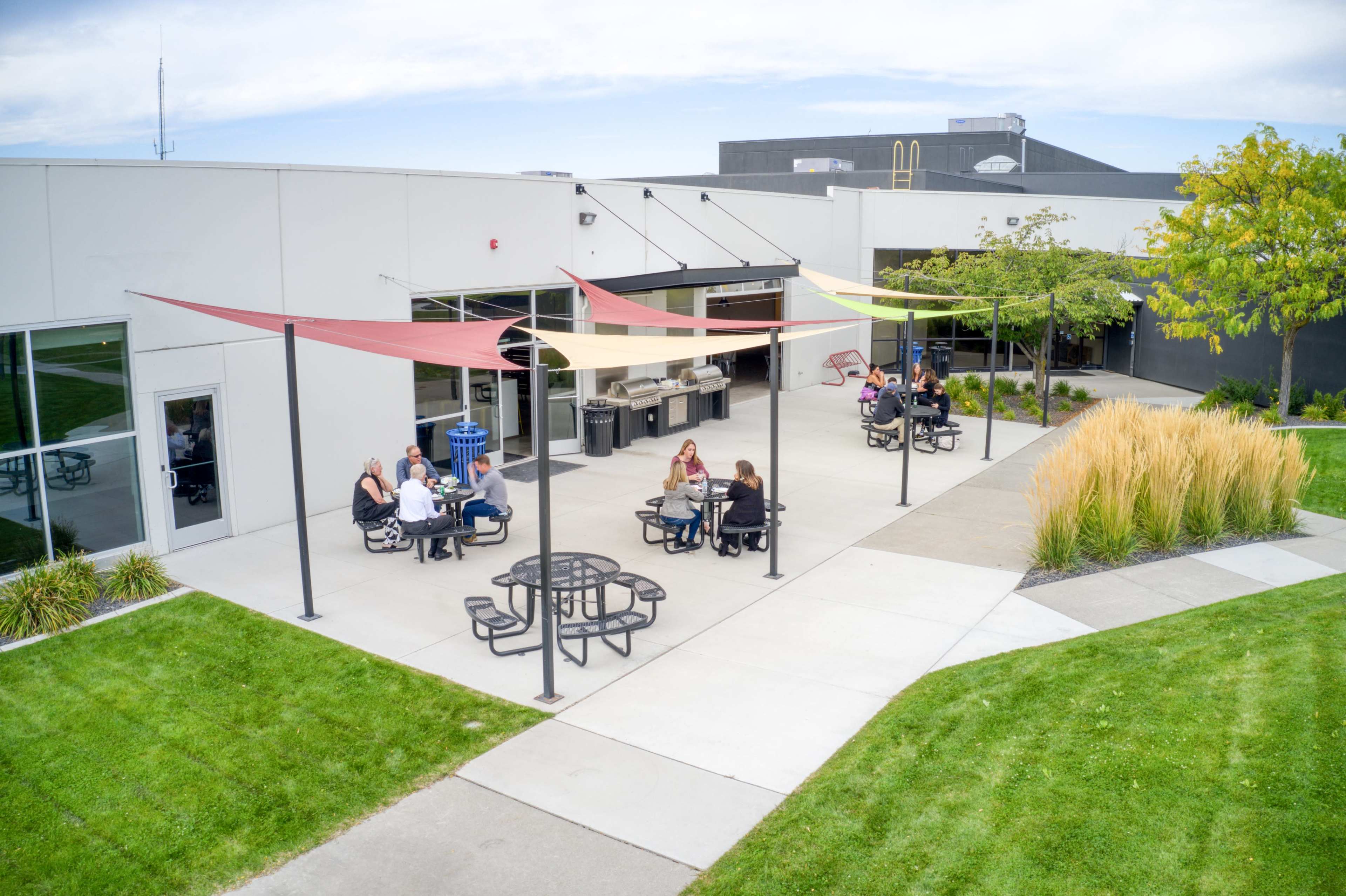 A group of people sits at tables under shaded canopies in a courtyard surrounded by grass and buildings.