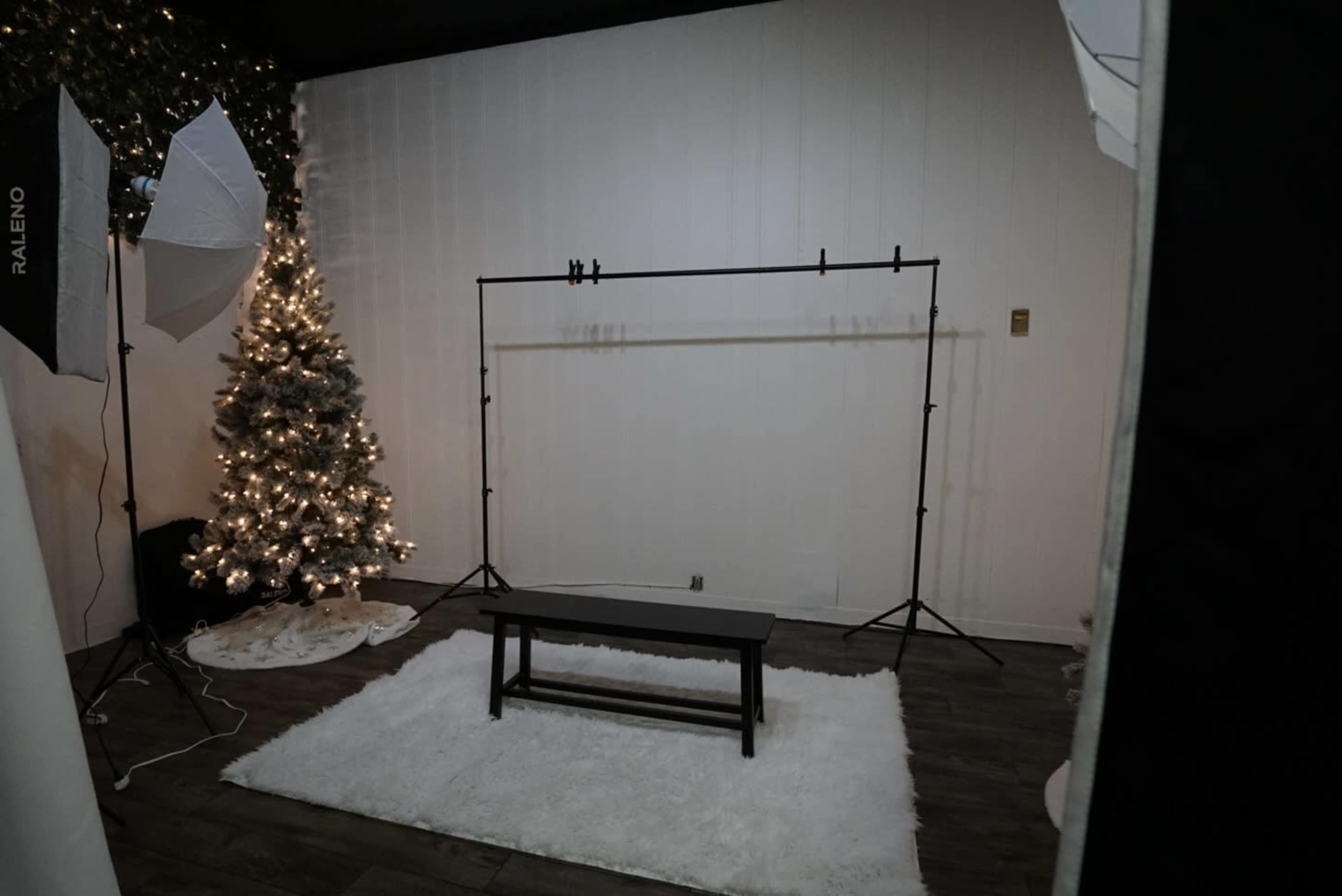 A photography studio setup with a black backdrop support system, a wooden bench on a white rug, and a decorated Christmas tree beside it.