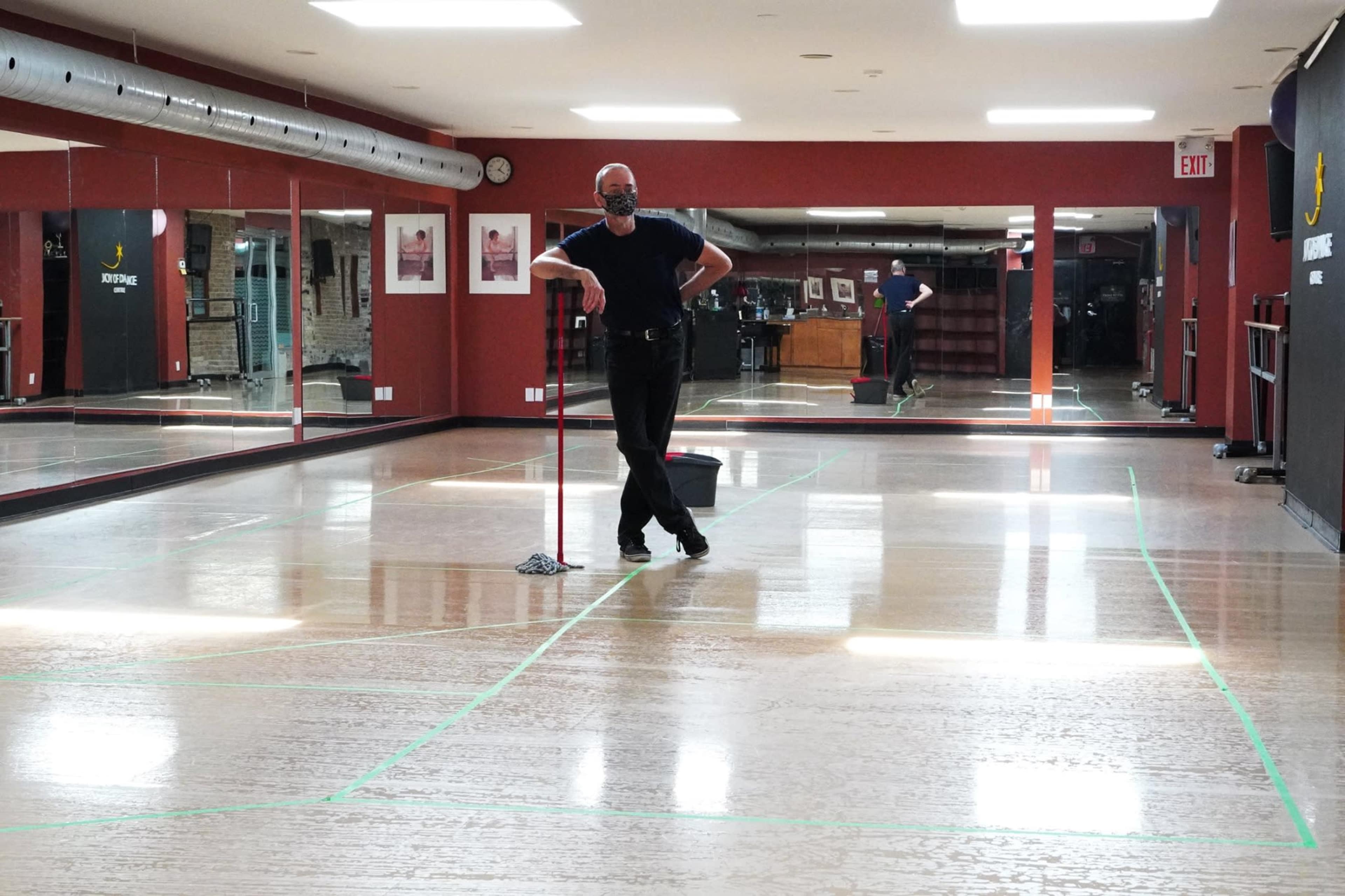 A man wearing a mask leans against a mop in a mirrored dance studio with green tape marking the floor.
