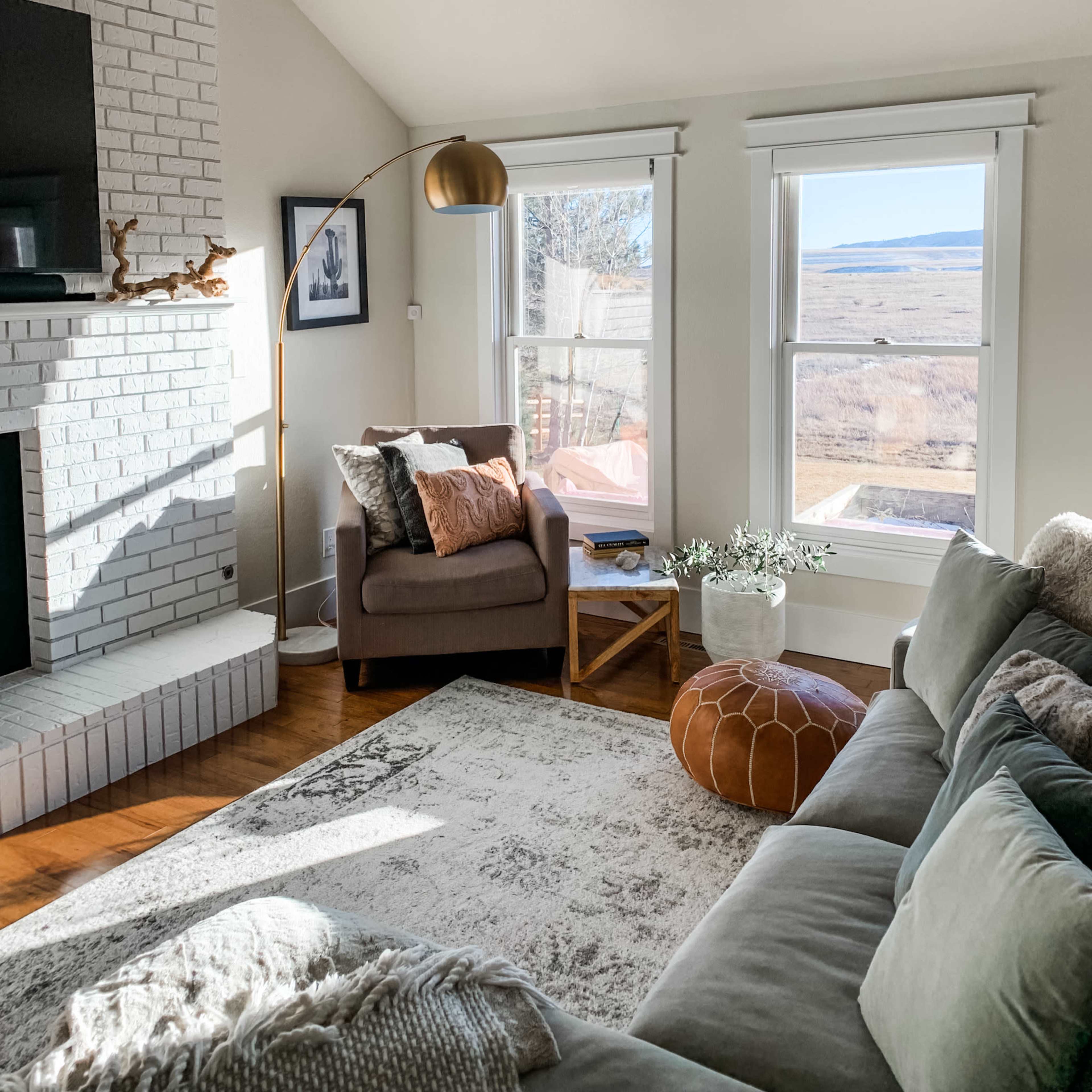A cozy living room features a green sofa, a brown armchair with decorative pillows, a wooden side table, and large windows overlooking a scenic view.