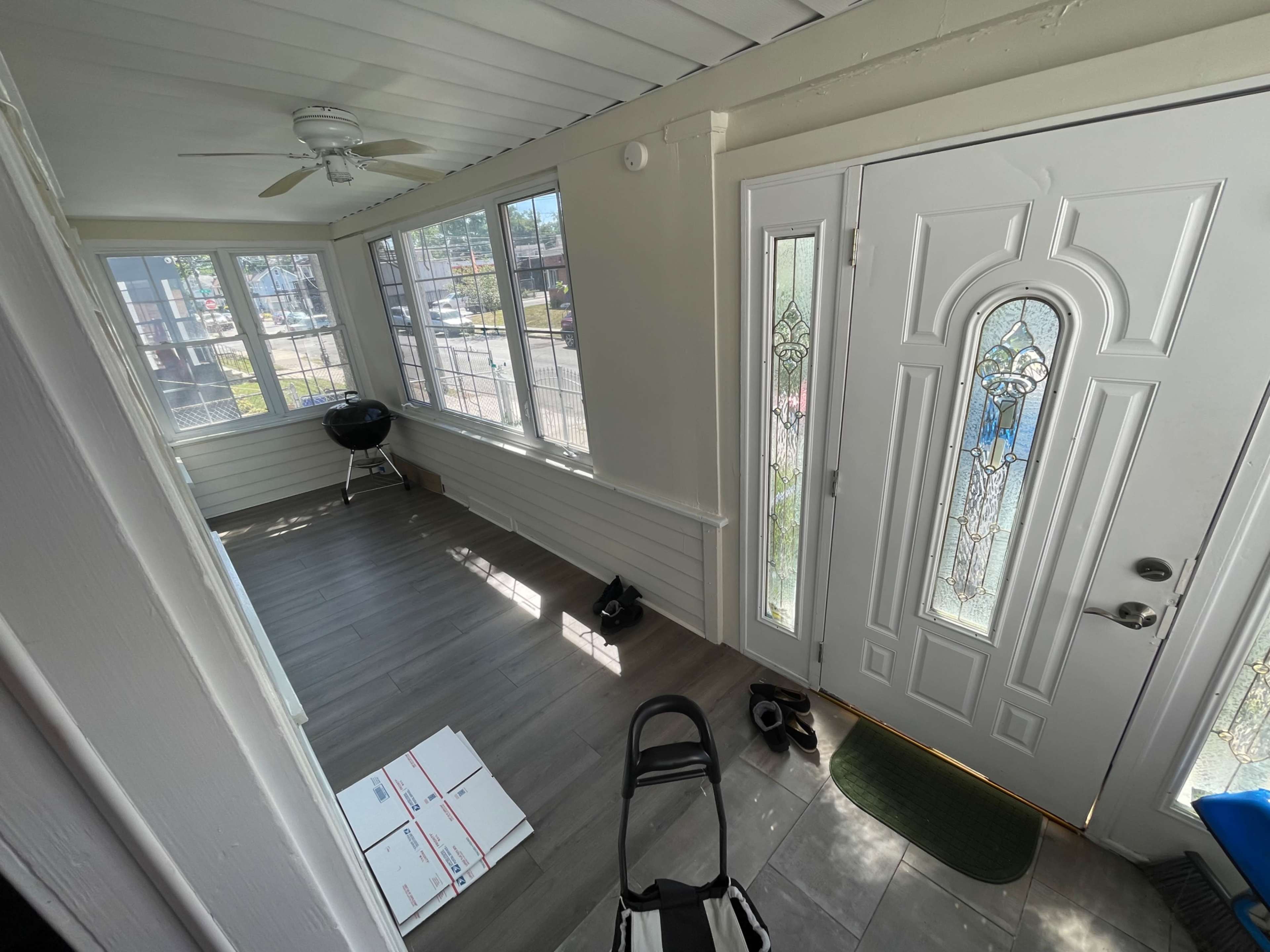 The image shows a bright entryway with large windows, a door with decorative glass, a small grill, and a few shoes on the floor.