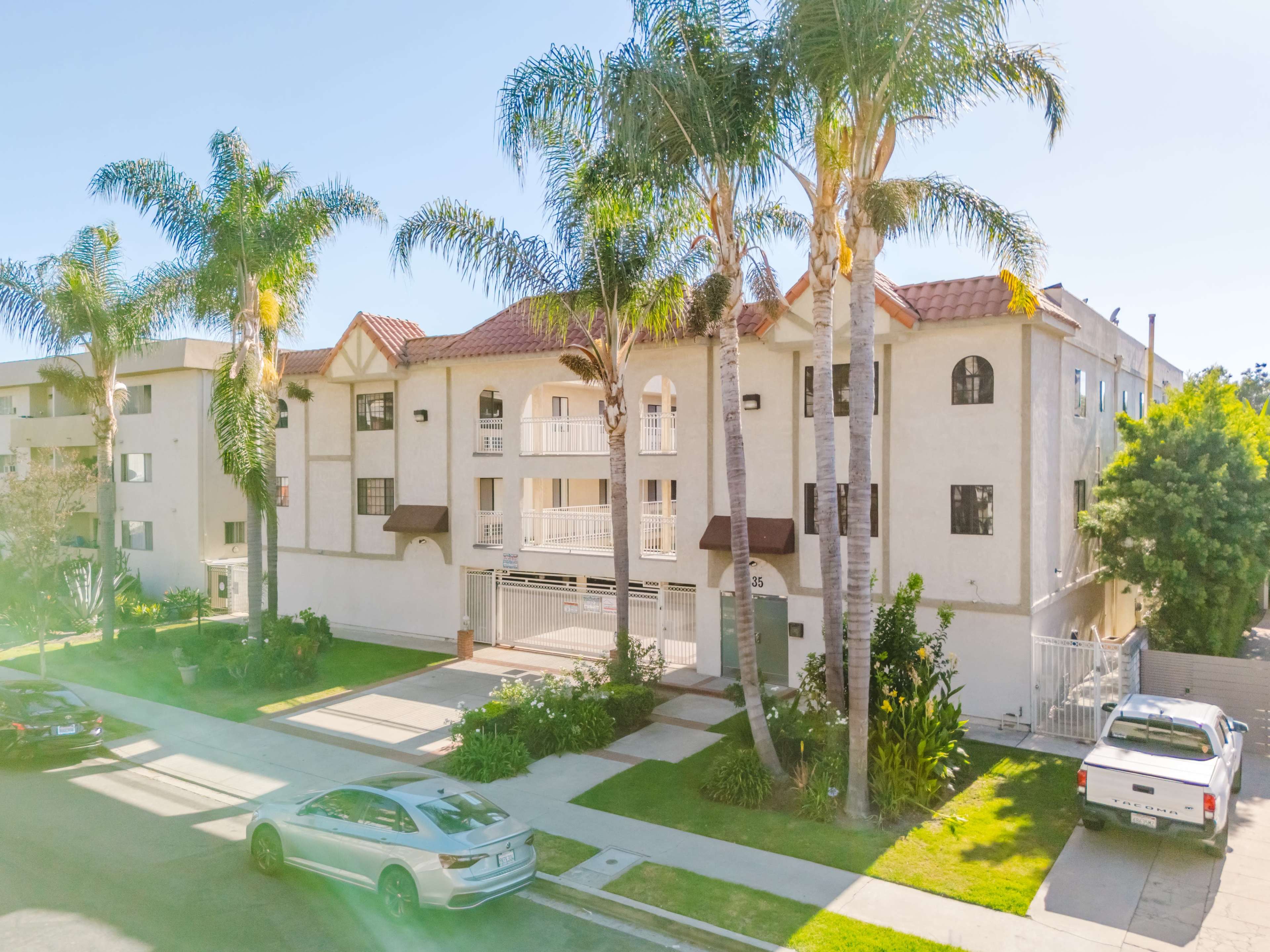 The image shows a multi-unit apartment building with palm trees in front and parked cars along the street.