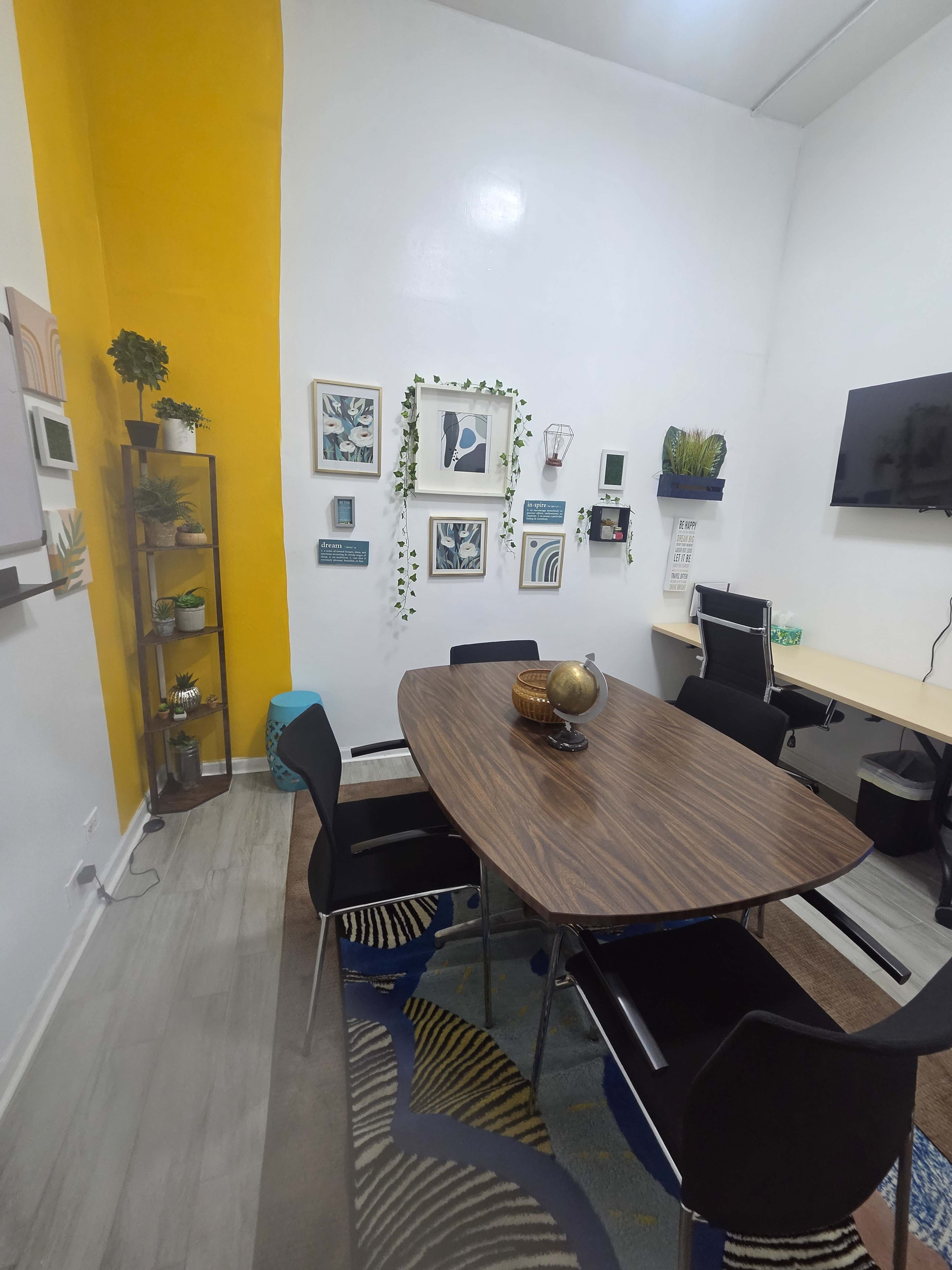 A modern meeting room features a wooden table surrounded by black chairs, with vibrant yellow and white walls adorned with framed artwork and plants.