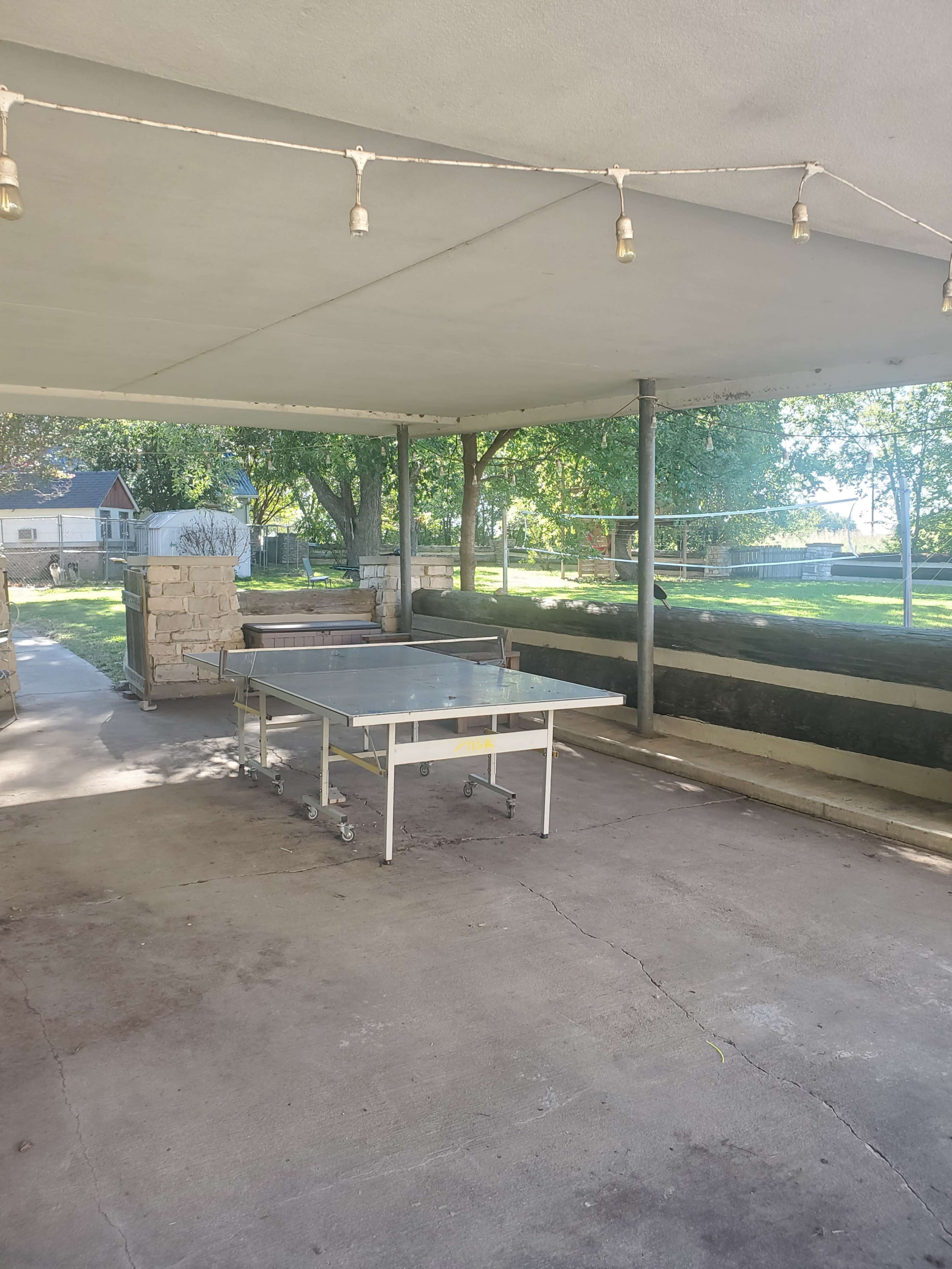 The image shows a covered outdoor area with a metal table on wheels and a stone structure along one side, surrounded by greenery.