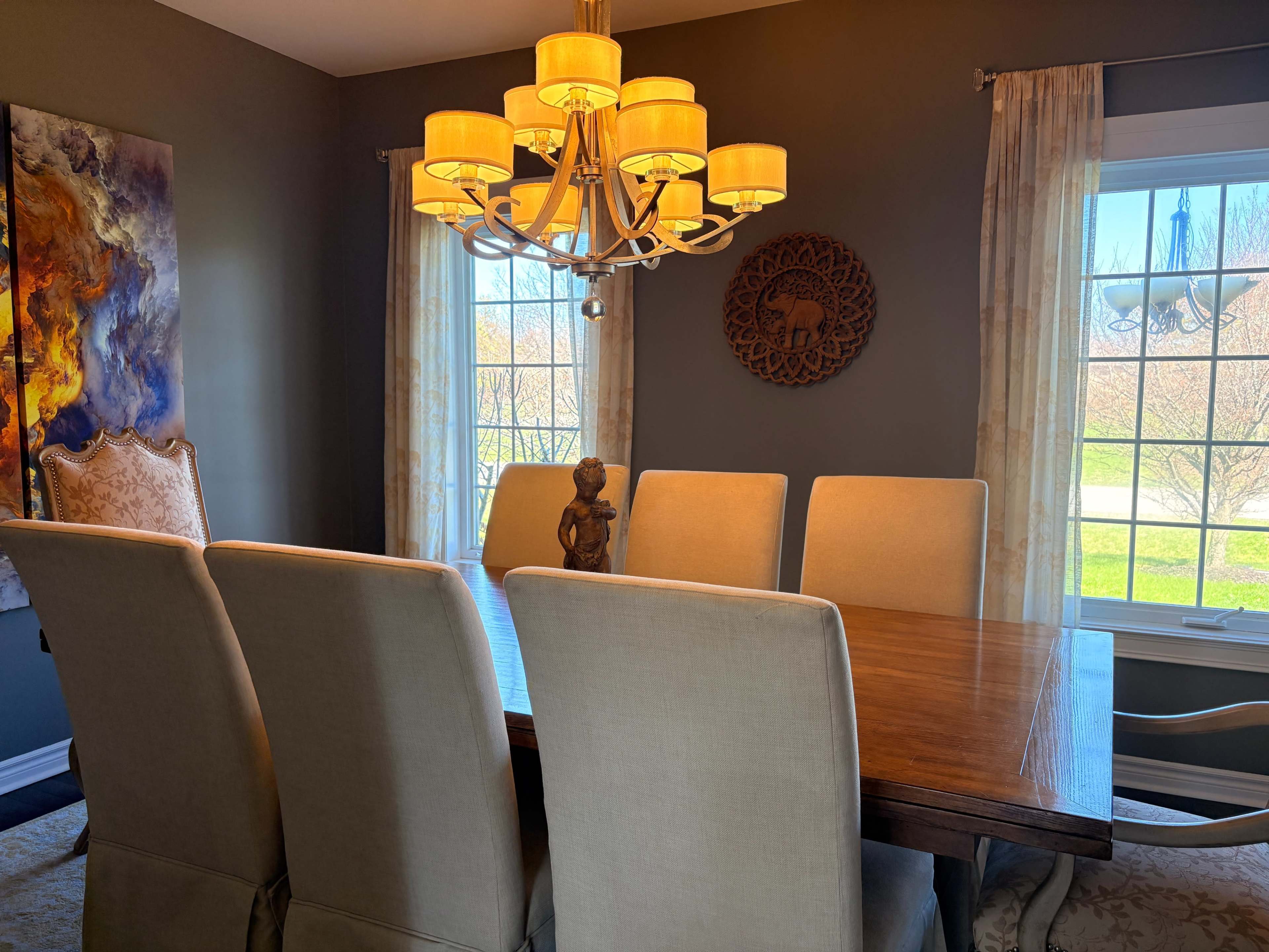 The image shows a dining room with a large wooden table surrounded by upholstered chairs, illuminated by a chandelier and flanked by windows with sheer curtains.
