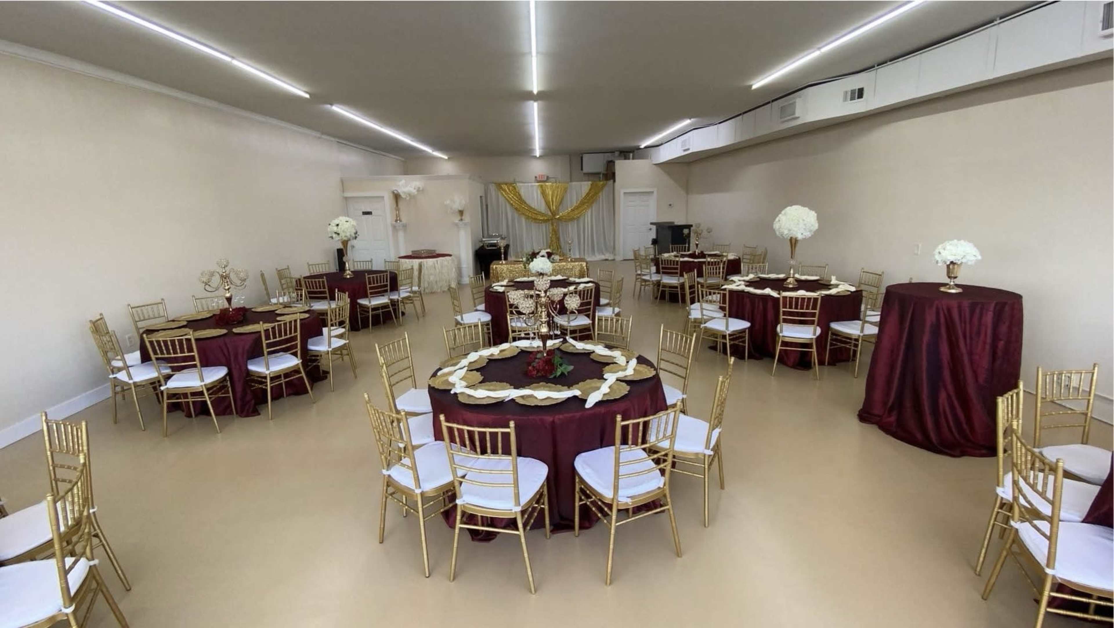 A banquet hall is set up with round and tall tables covered in burgundy tablecloths, gold chairs, and floral centerpieces.