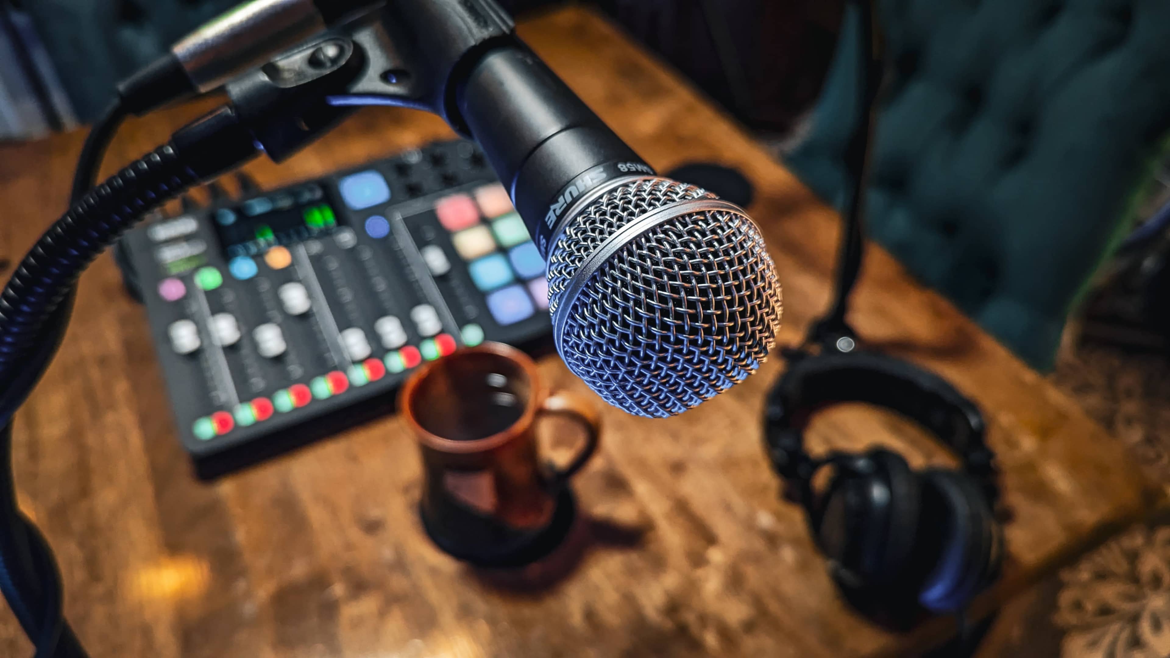 A microphone is positioned in front of a mixing board, with a coffee cup and headphones visible on a wooden table.