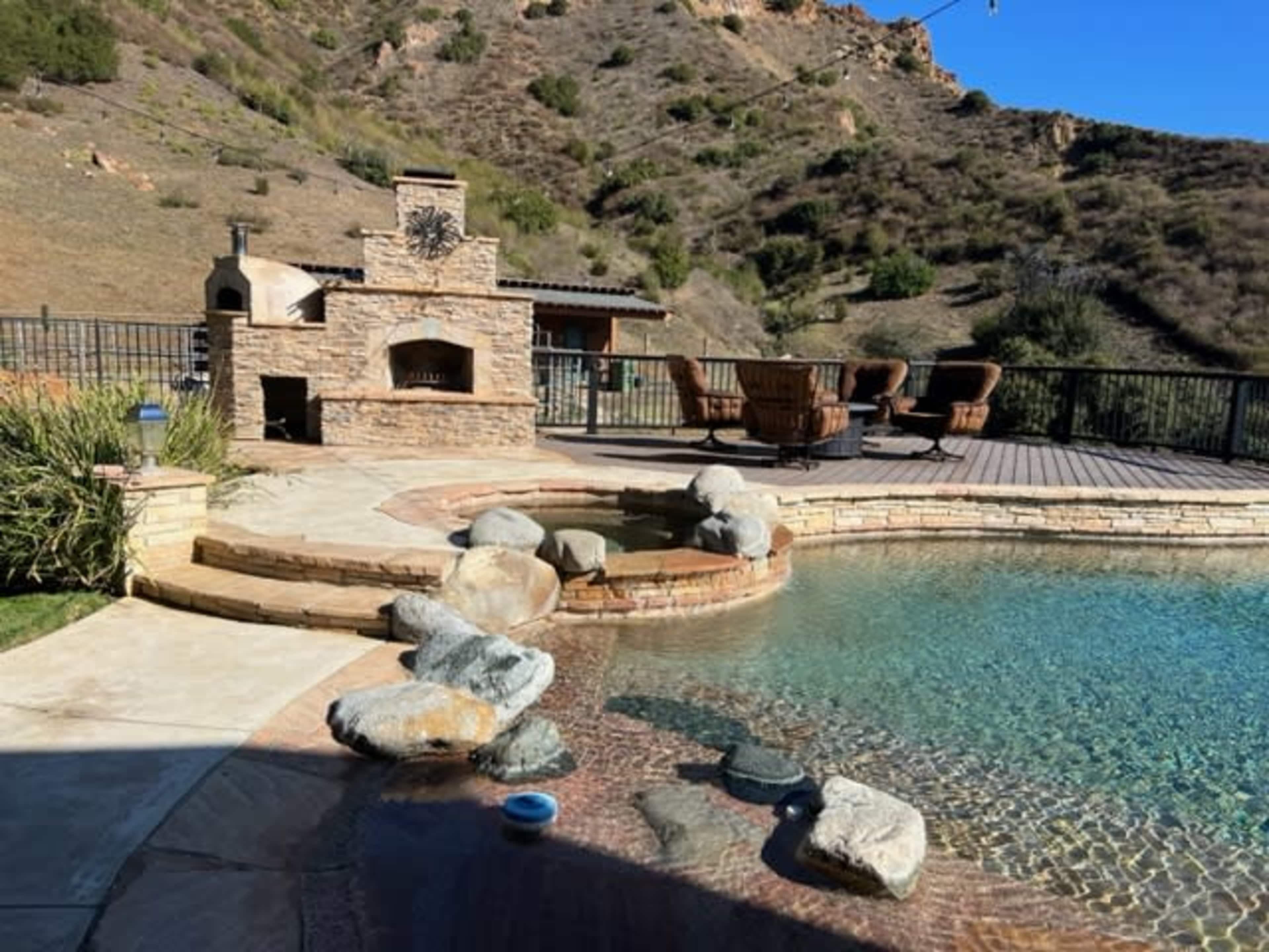 A stone patio with a swimming pool, a built-in outdoor grill, and a view of a hillside in the background.