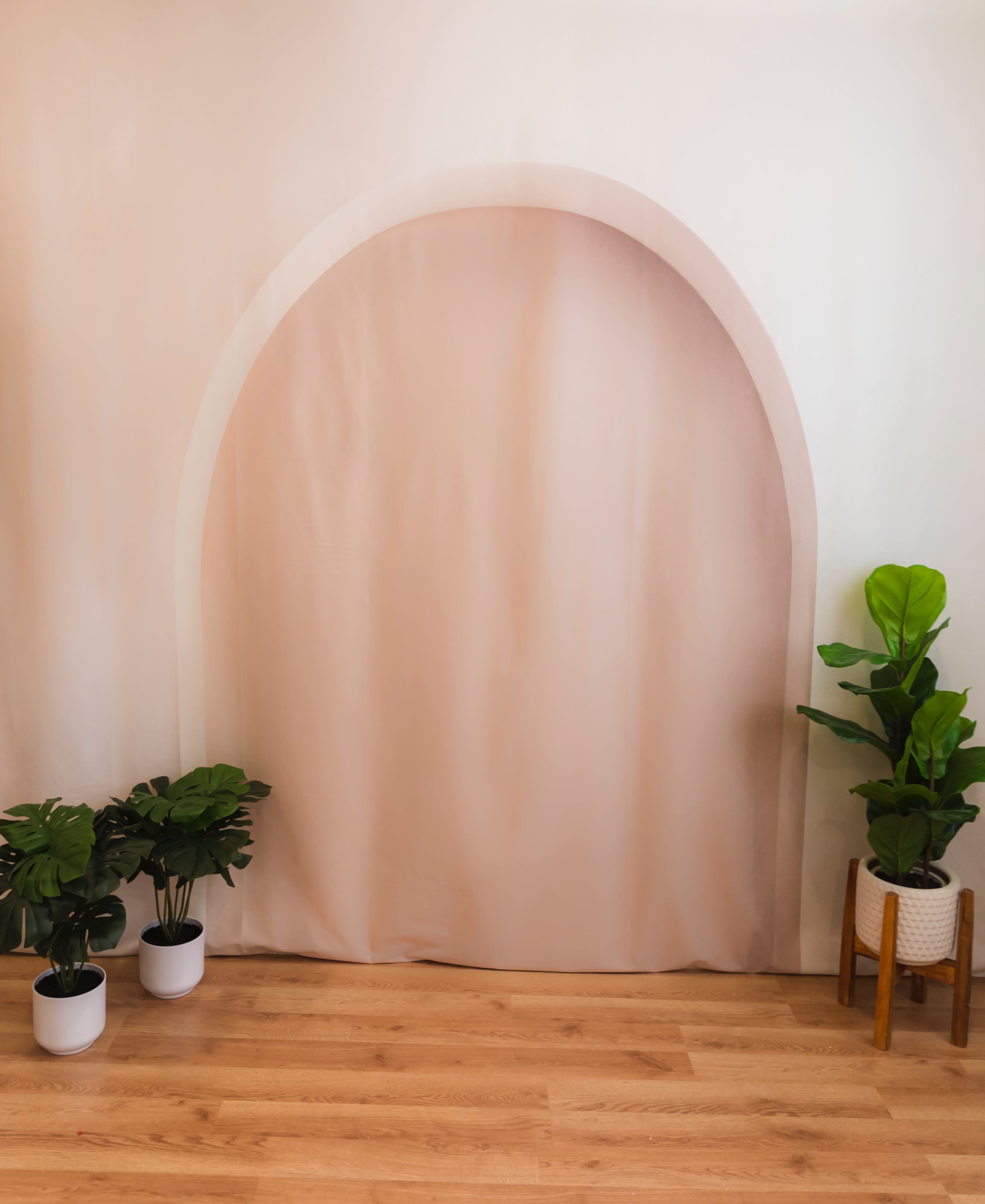 The image features a soft pink backdrop with an arch design, flanked by two potted plants on the floor.
