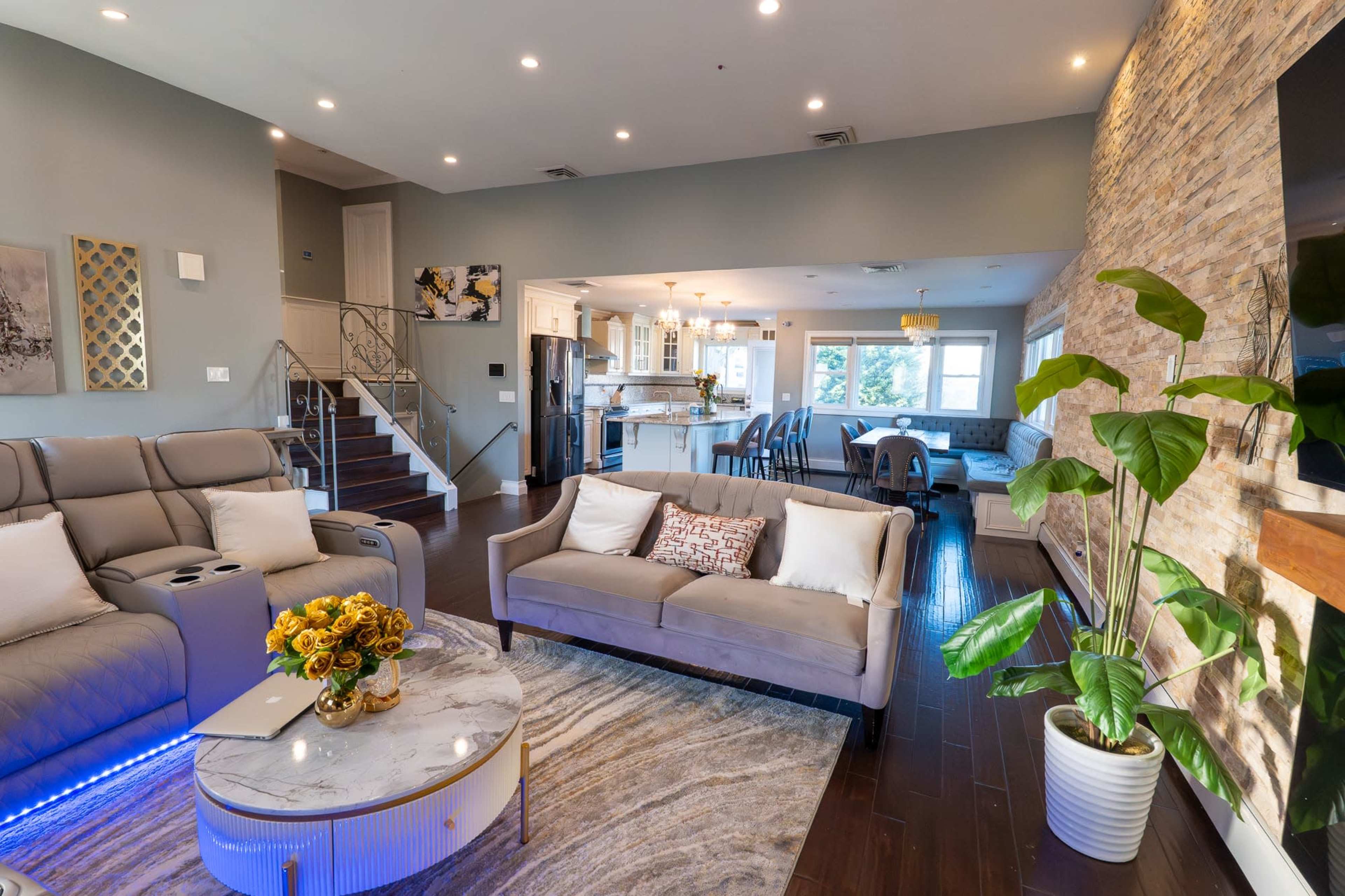 The image shows a modern living room with a sectional sofa, a coffee table, and a staircase leading to another level, complemented by a kitchen area in the background.