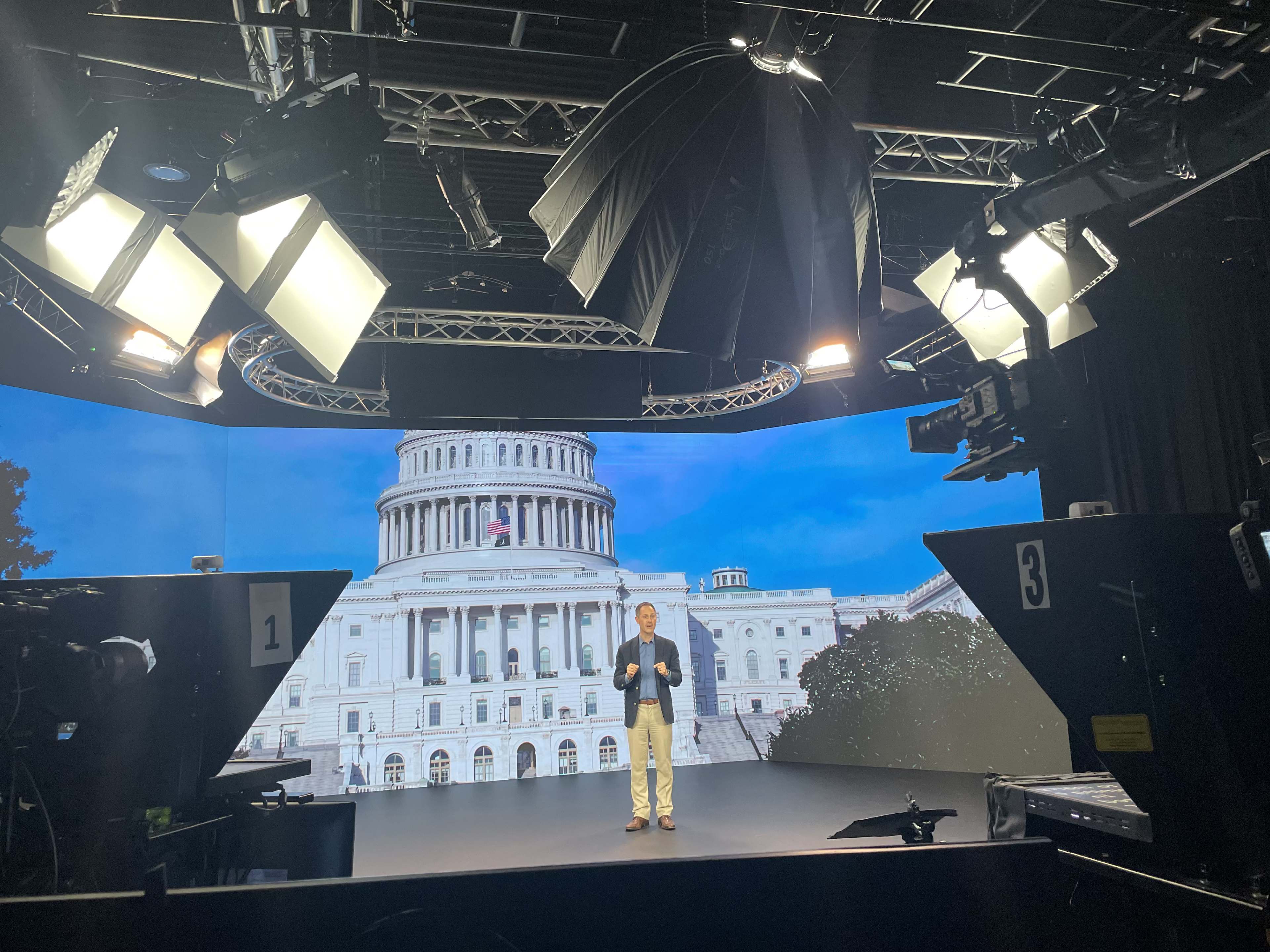 A man stands on a stage in a studio, with large lights overhead and a backdrop displaying the U.S. Capitol building.