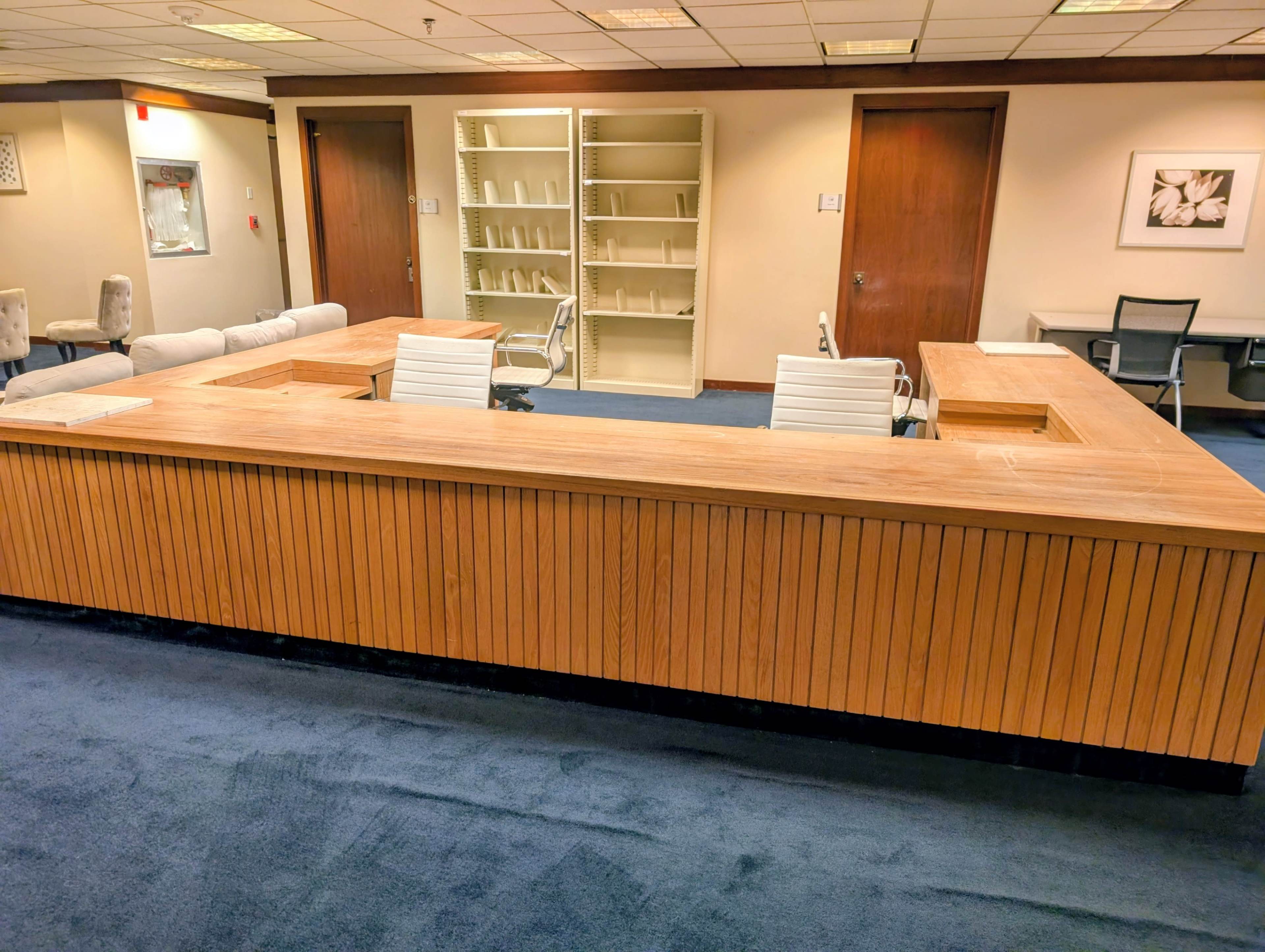 The image shows a wooden reception desk in an office space with empty shelves and a few chairs arranged nearby.