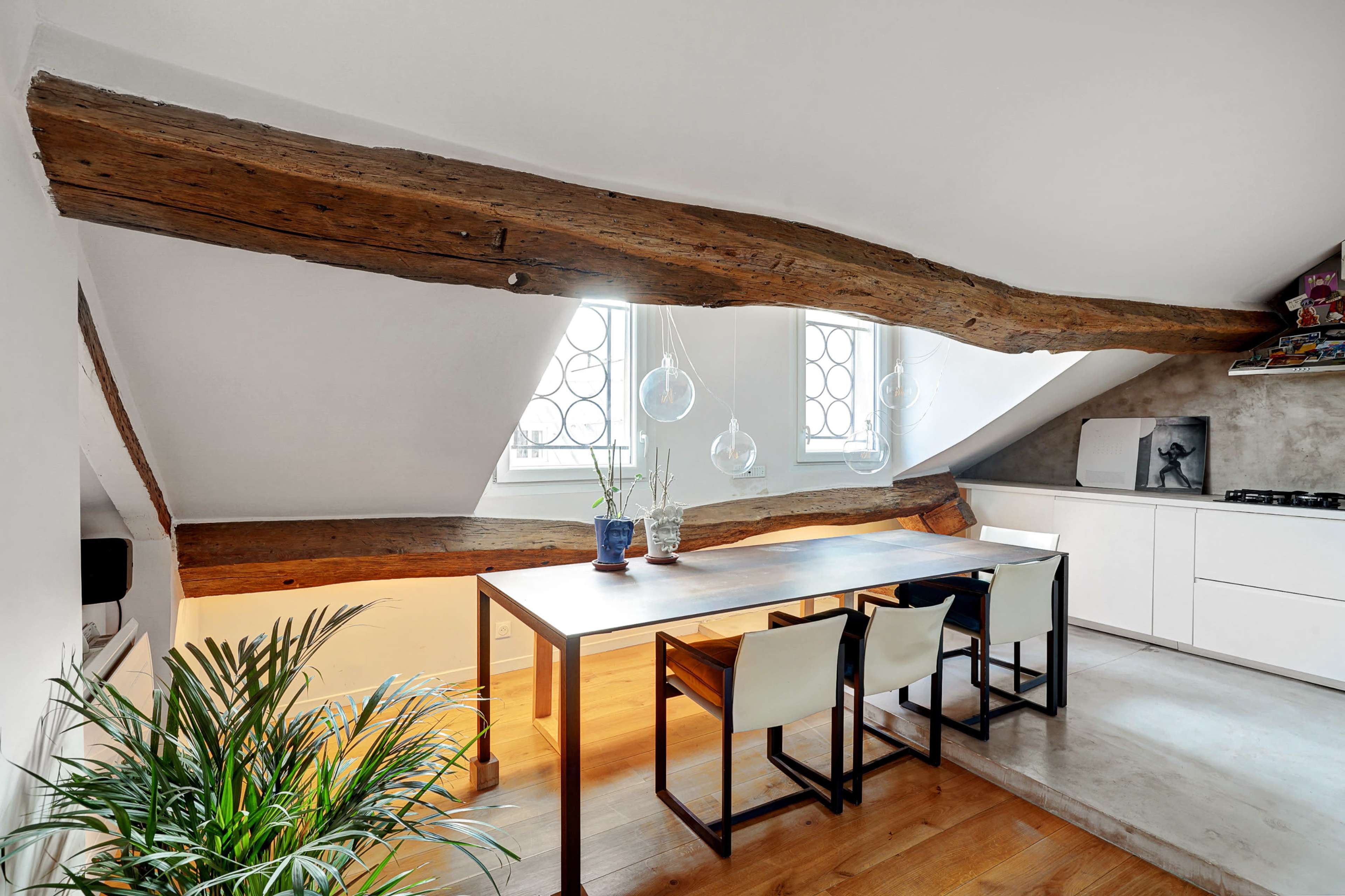 A modern attic dining area with a wooden table, four chairs, large windows, and exposed wooden beams.