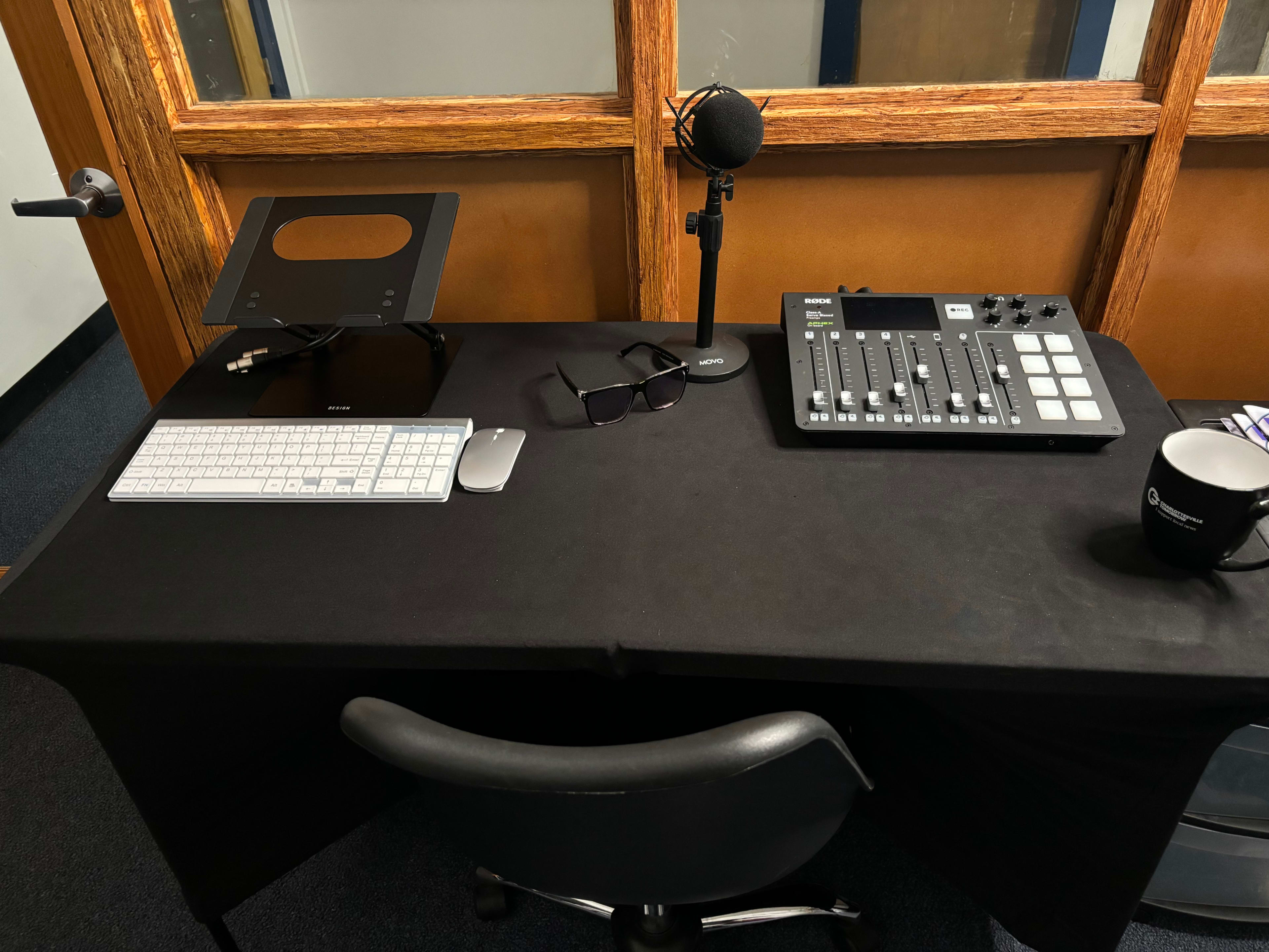 A desk setup with a keyboard, microphone, audio mixer, glasses, and a coffee mug on a black tablecloth.