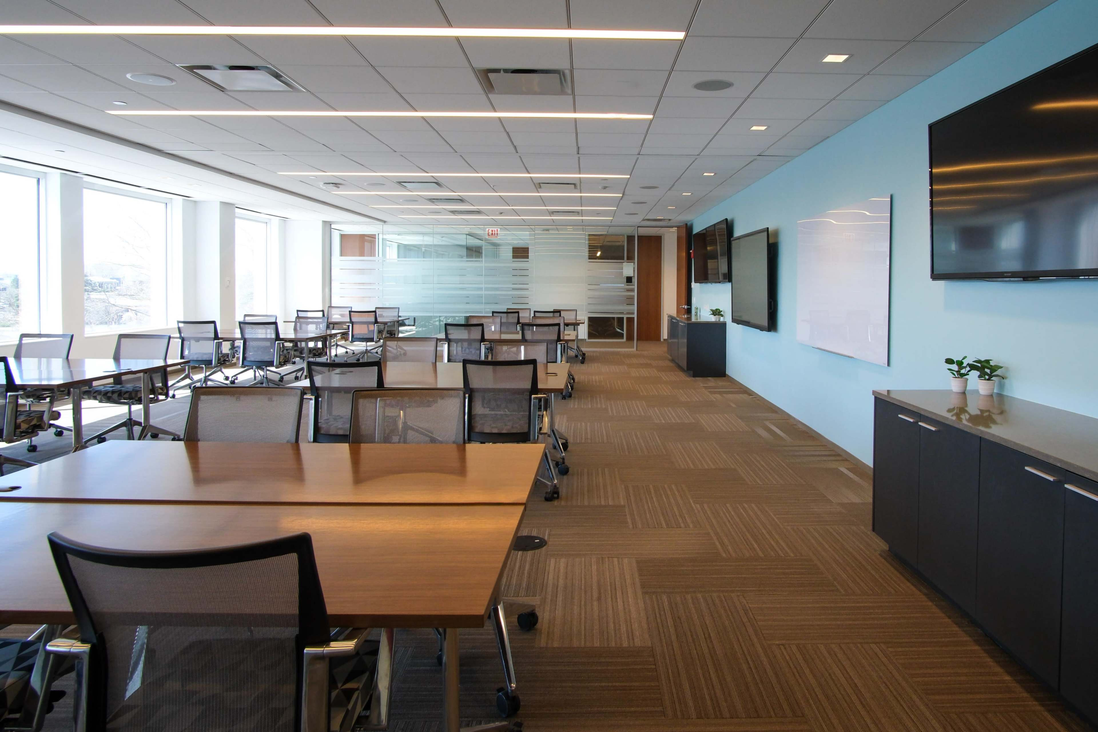 A modern office conference room features multiple tables and chairs arranged in a spacious layout with large windows and a blue accent wall.