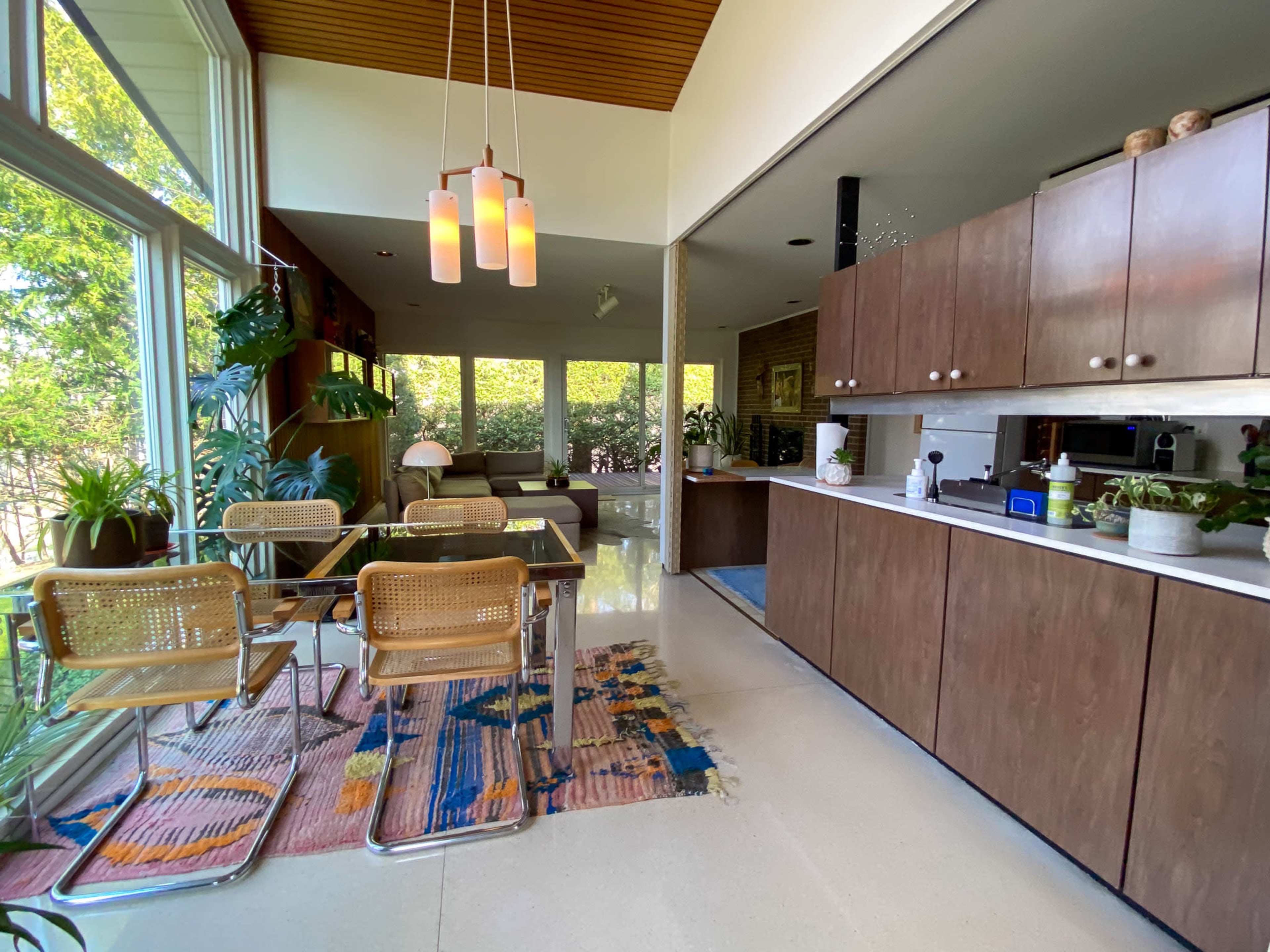 The image shows a modern kitchen and dining area with wooden cabinetry, a glass table surrounded by chairs, and large windows allowing natural light to fill the space.