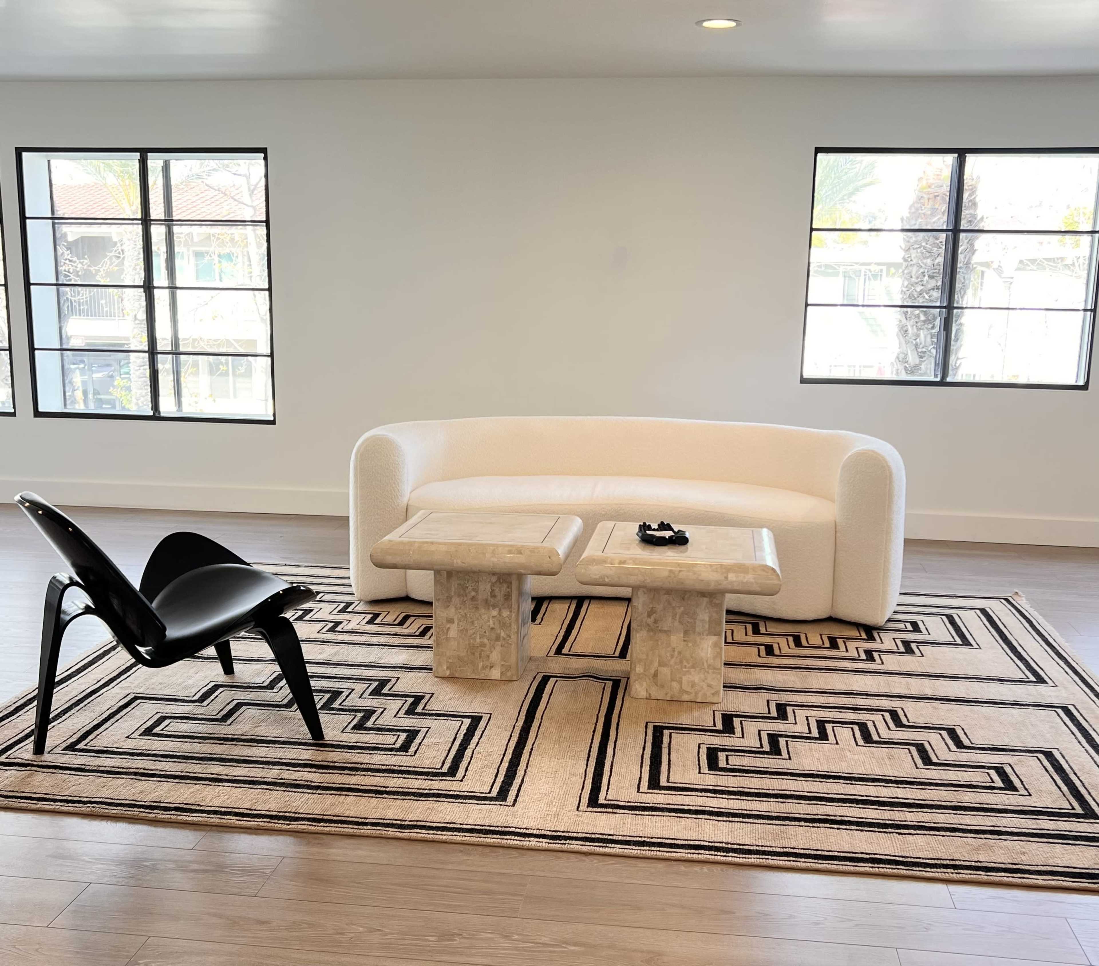A modern living area featuring a white curved sofa, a pair of stone tables, a black chair, and a geometric-patterned rug, with large windows allowing natural light.