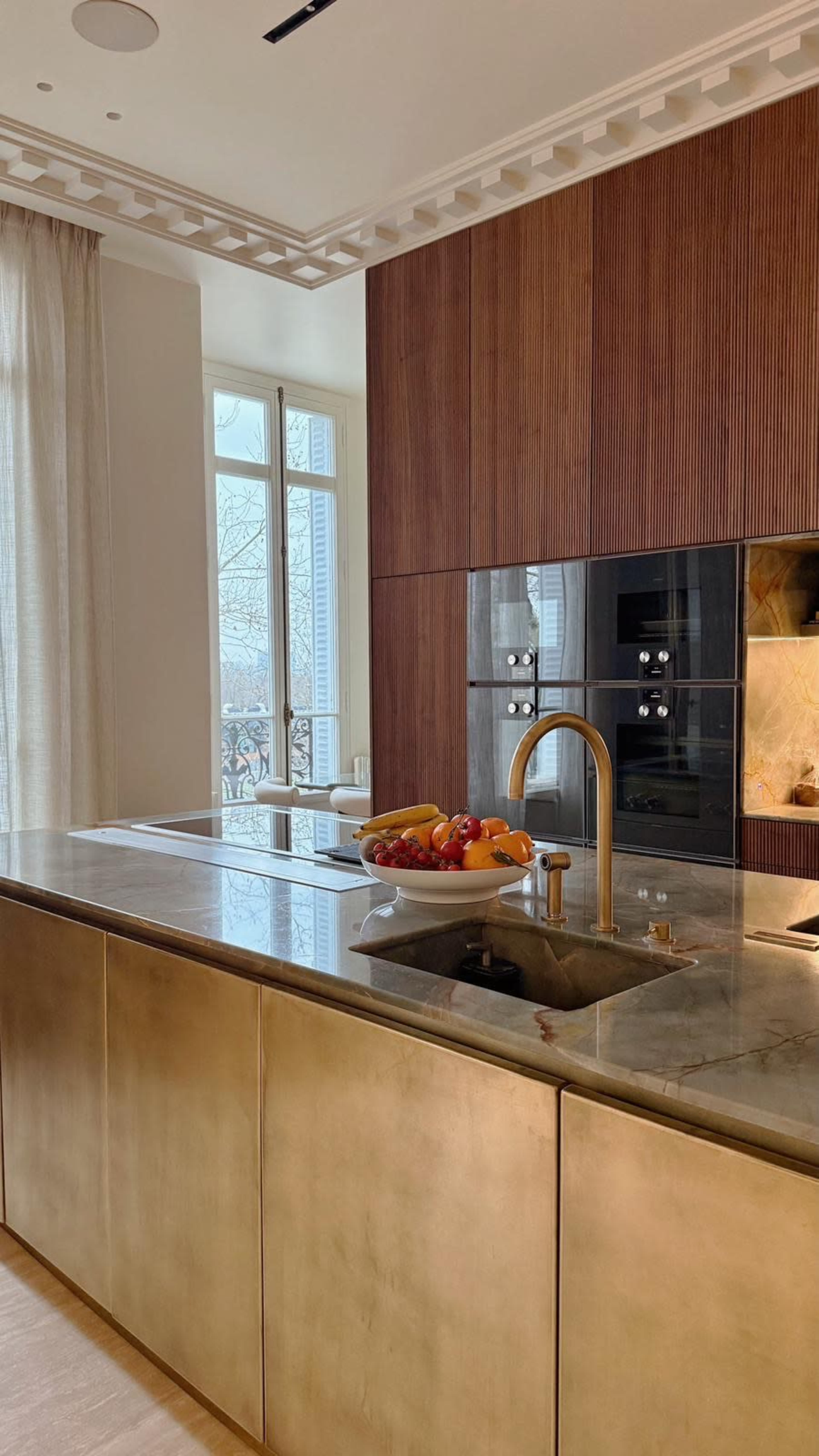 A modern kitchen features a brass island with a sink and a bowl of fruit, alongside wooden cabinetry and large windows allowing natural light.