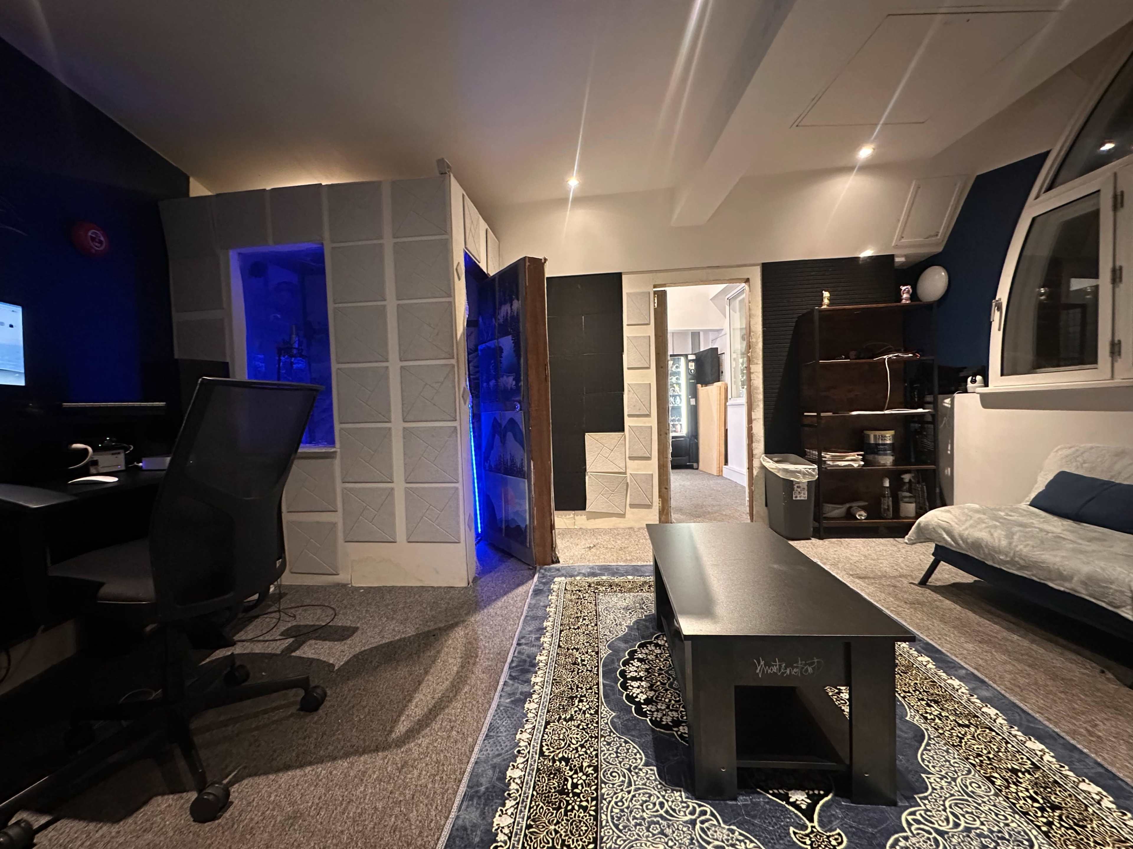 The image shows a modern studio room featuring a desk with a computer, a patterned rug, a dark table, and partitions creating separate spaces.