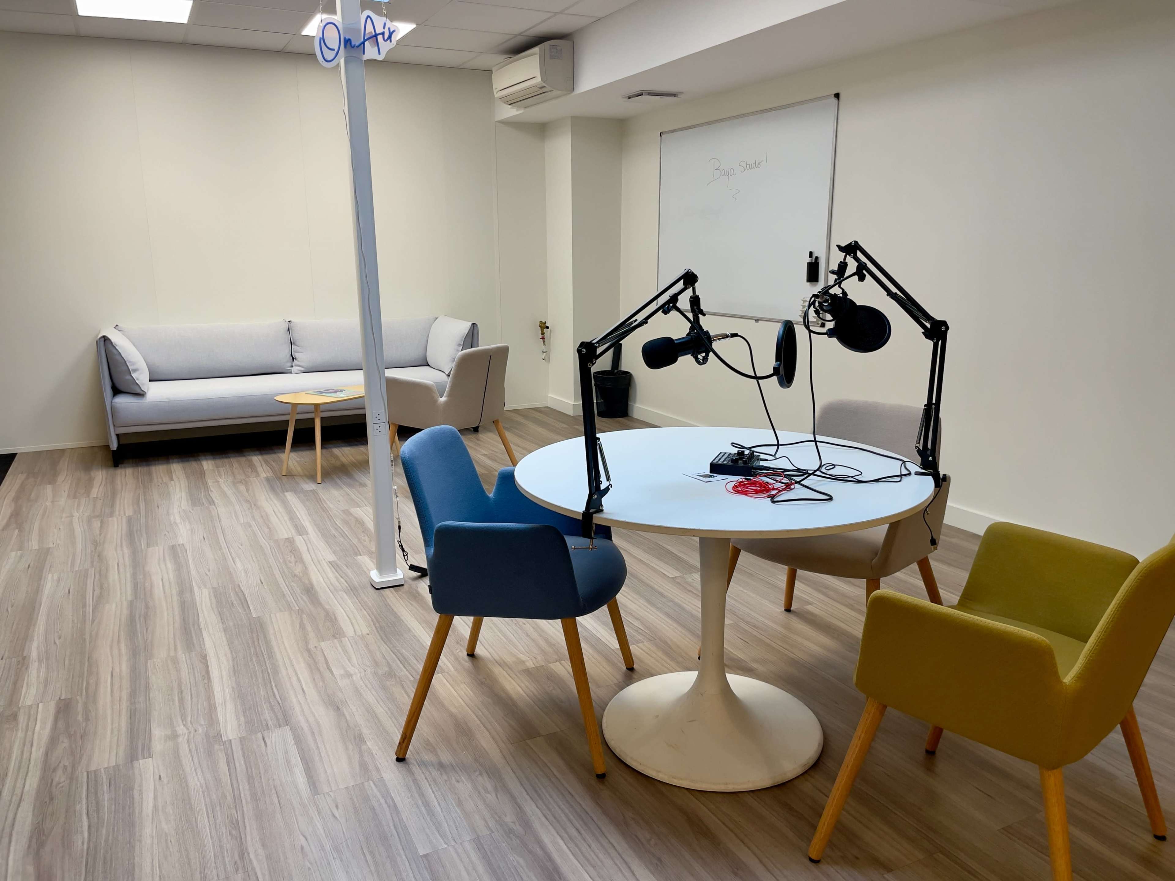 The image shows a modern recording studio with a round table, colorful chairs, microphones, and a sofa in a well-lit room.
