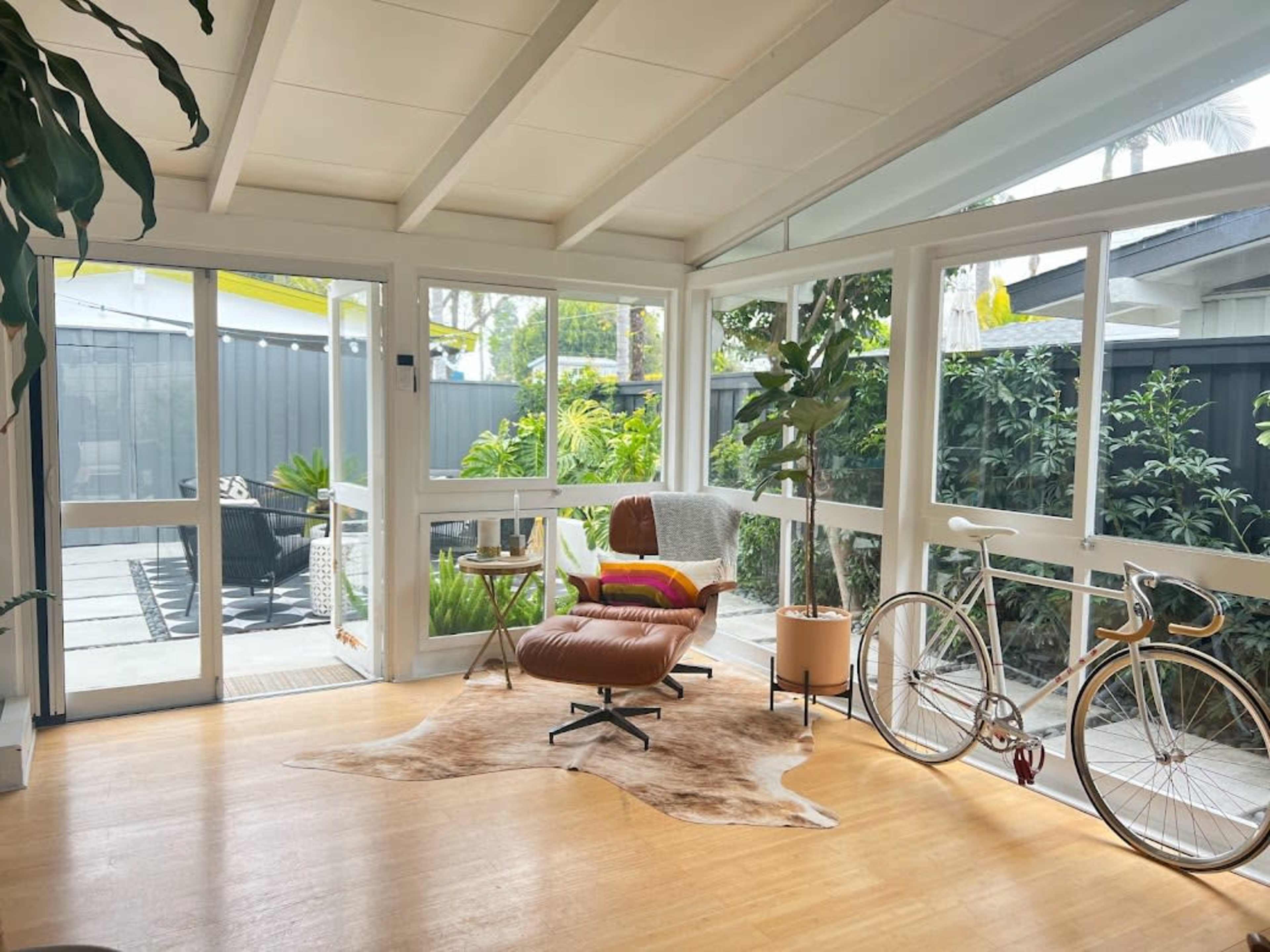 A bright sunroom features a brown leather chair, a bicycle, and plants, with glass walls opening to an outdoor area.
