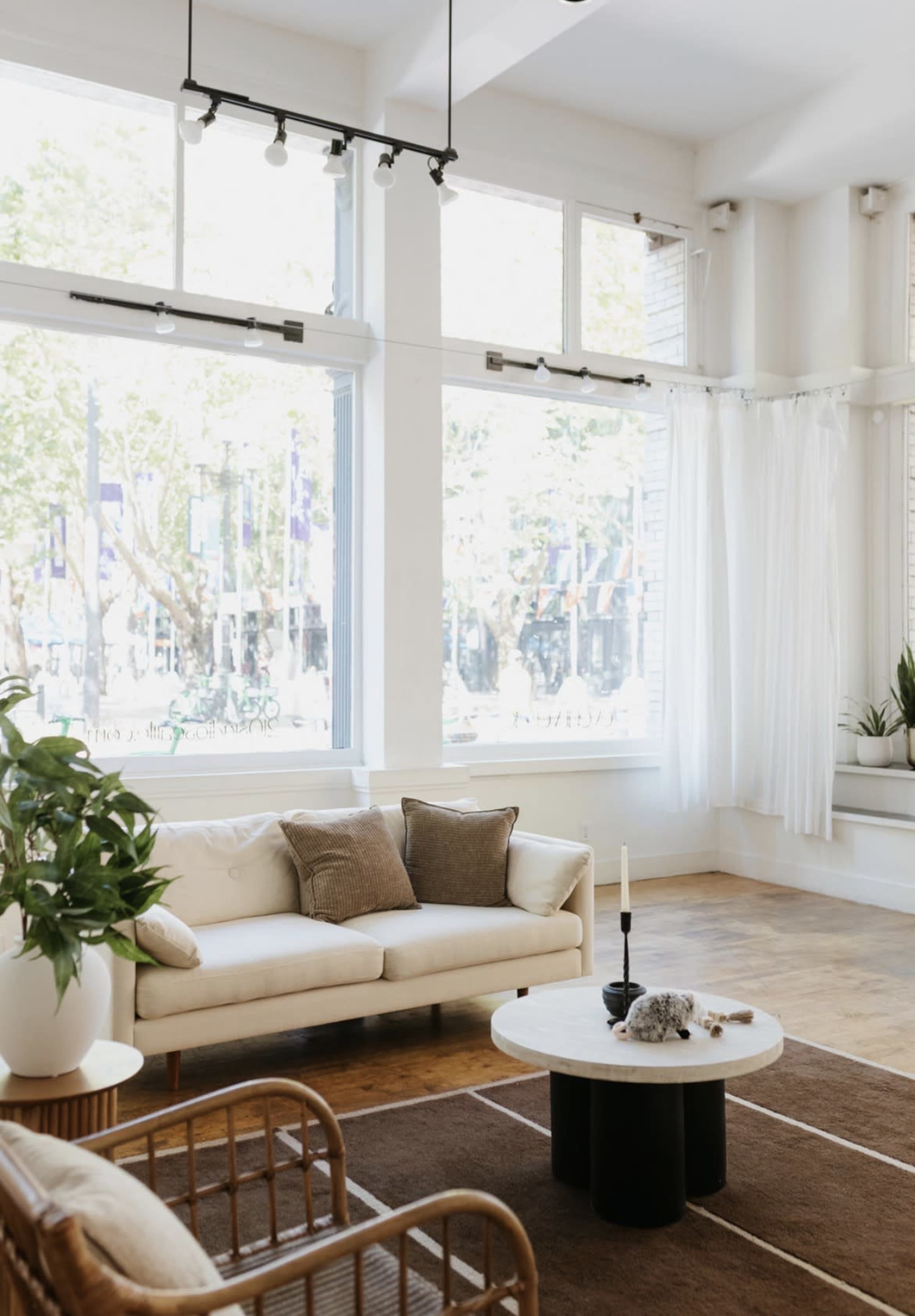 The image shows a bright and airy living room with large windows, a beige sofa adorned with cushions, a round coffee table, and a plant in the corner.