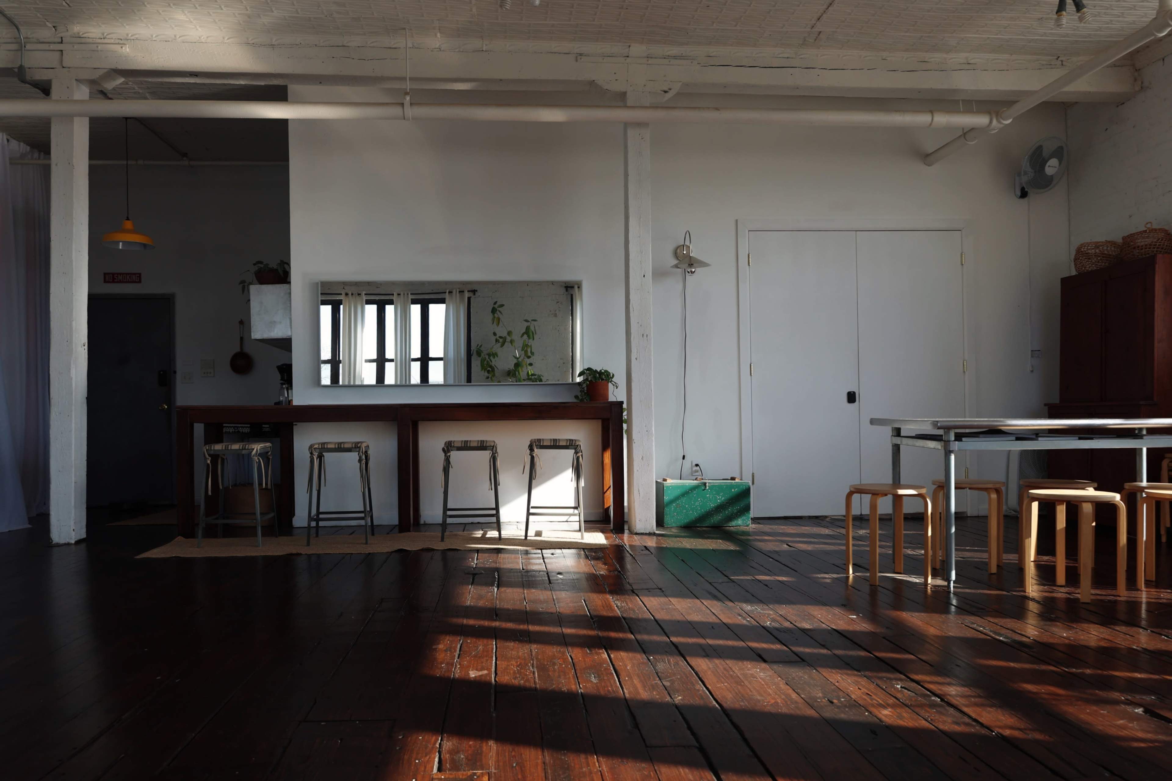 A spacious, airy interior features a long wooden bar with metal stools, a large mirror, and a dining table set against a backdrop of white walls and wooden flooring.